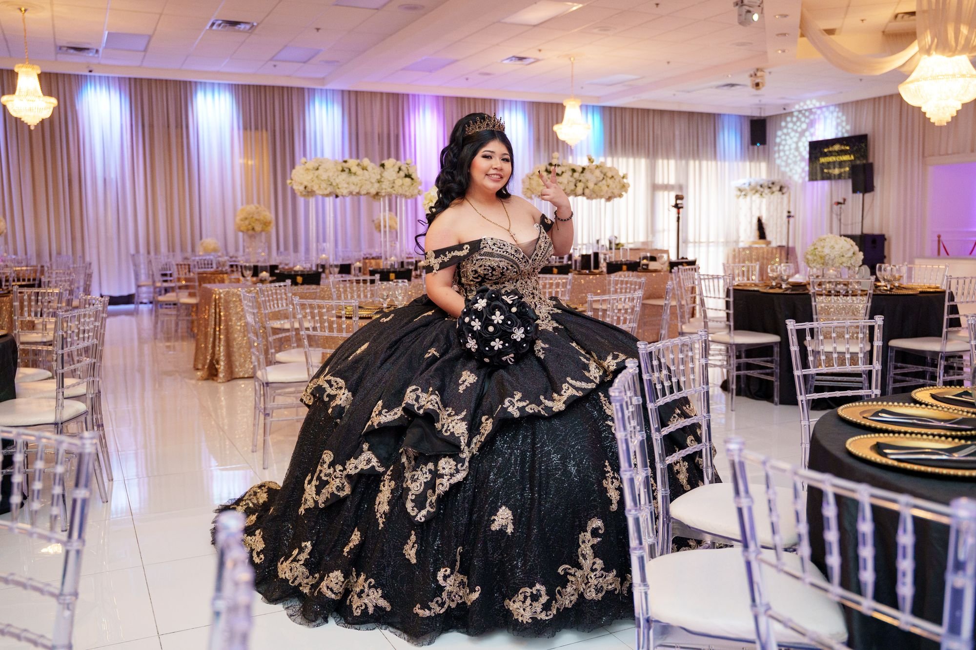 Quinceañera Jayden in black and gold ballgown with crown holding bouquet posing alone in empty Luxor Banquet Hall ballroom before guests arrive at her reception