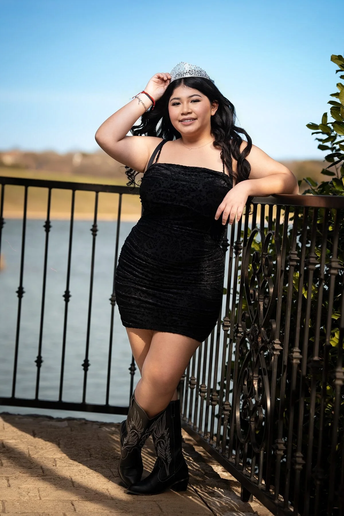 Jayden in black velvet dress and cowboy boots adjusts crystal tiara at lakefront wrought iron fence, Adriatica Village McKinney