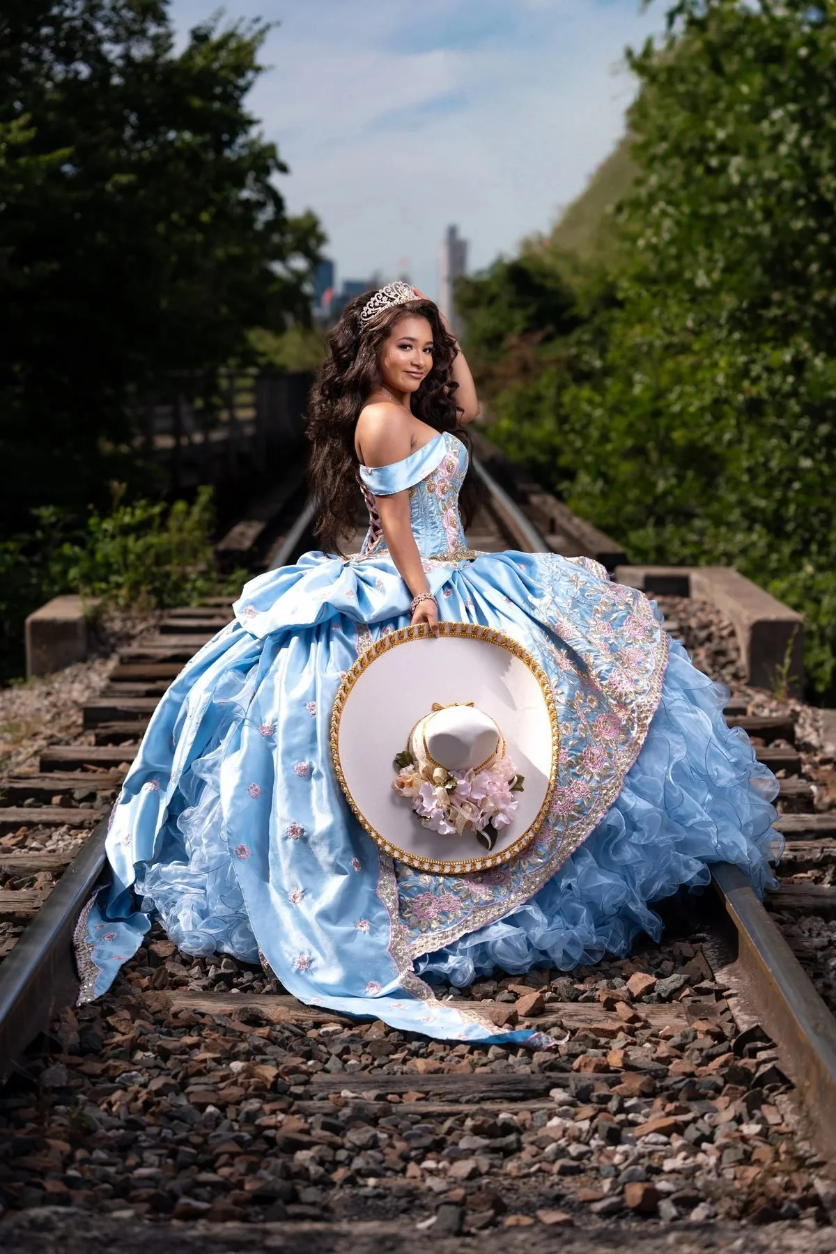 Quinceañera in a light blue charro ball gown holding a mariachi sombrero on railroad tracks in Fort Worth.