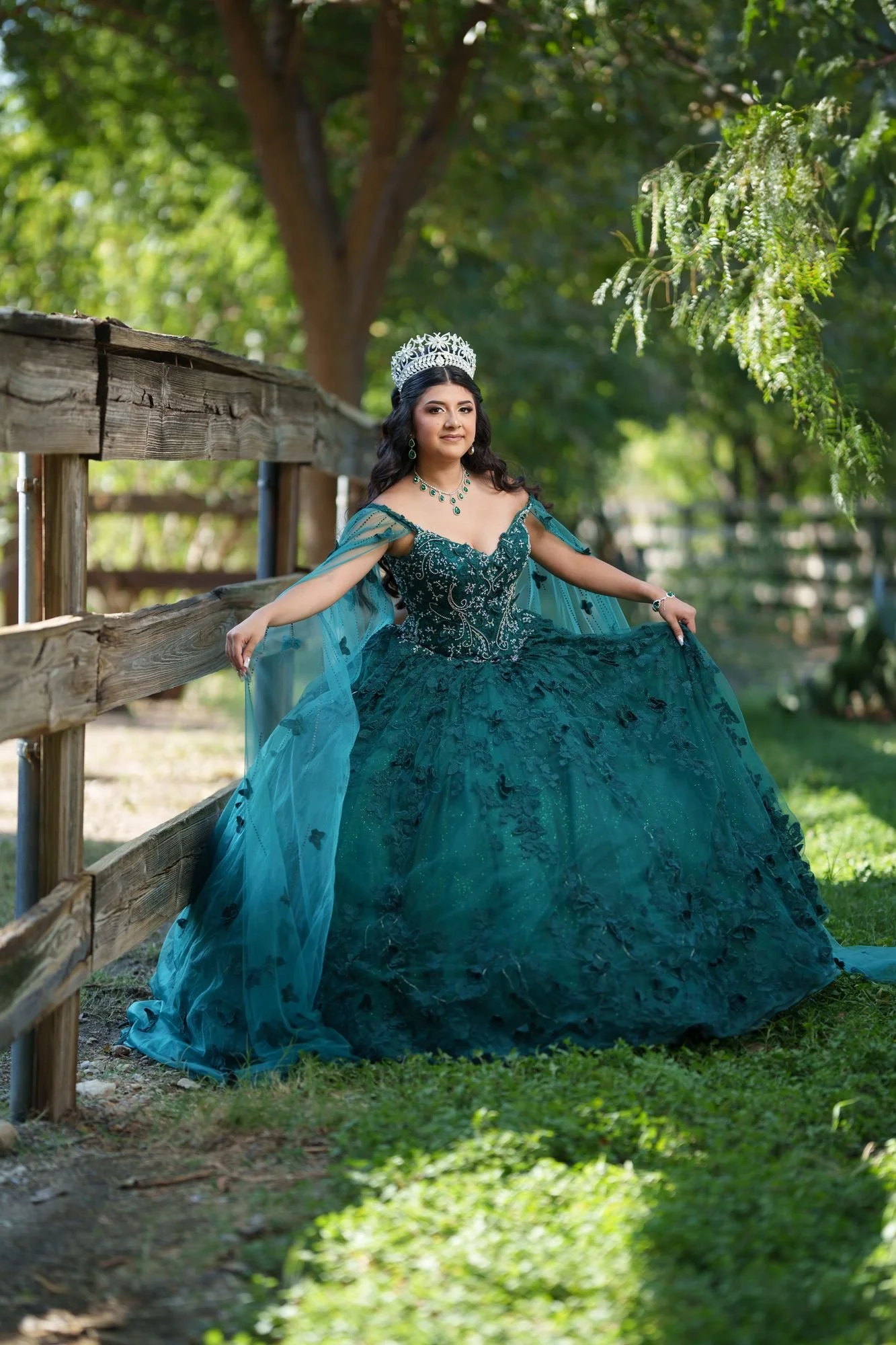 Nevaeh in her emerald quinceañera ballgown holding the dress out to the sides at a wooden fence with trees behind her during her Fort Worth Stockyards formal preshoot