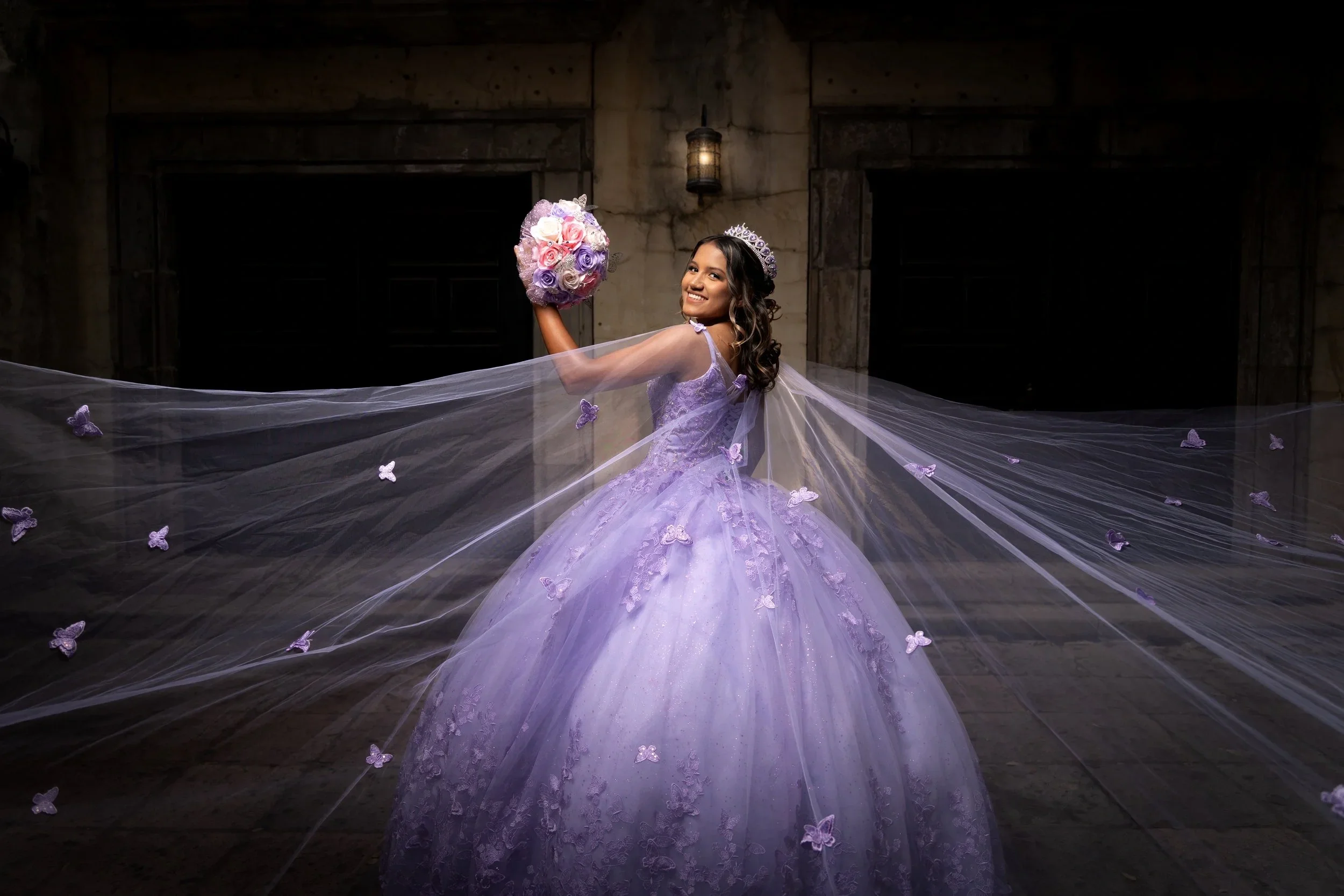 Quinceañera in a lavender gown with a flowing butterfly cape holding a bouquet against a stone castle backdrop in DFW