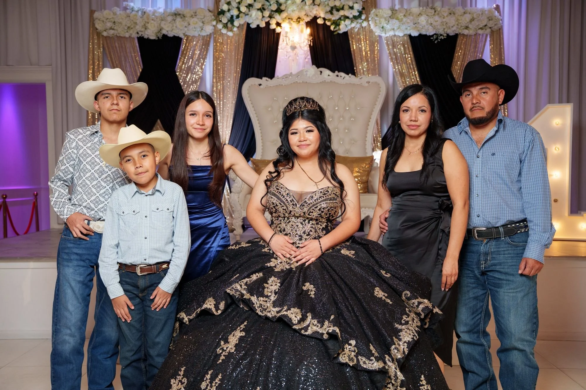 Jayden in black and gold ballgown seated on white throne with extended family in coordinated Western attire including cowboy hats and boots at Luxor Banquet Hall Carrollton TX quinceañera