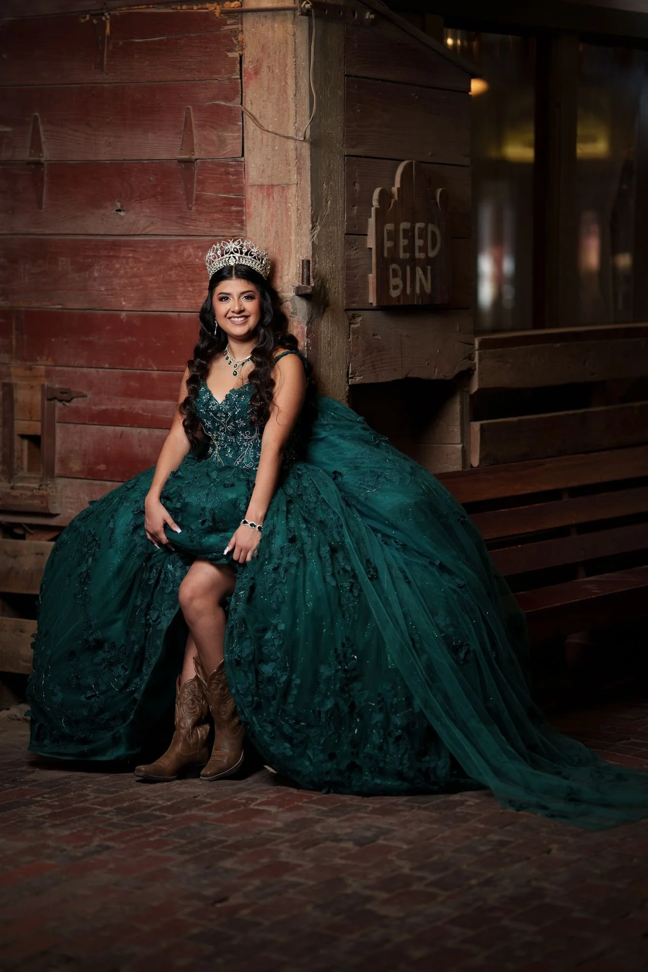 Nevaeh smiling directly at the camera in her emerald quinceañera ballgown and silver tiara beneath the historic Feed Bin sign at Fort Worth Stockyards, with brown cowboy boots visible