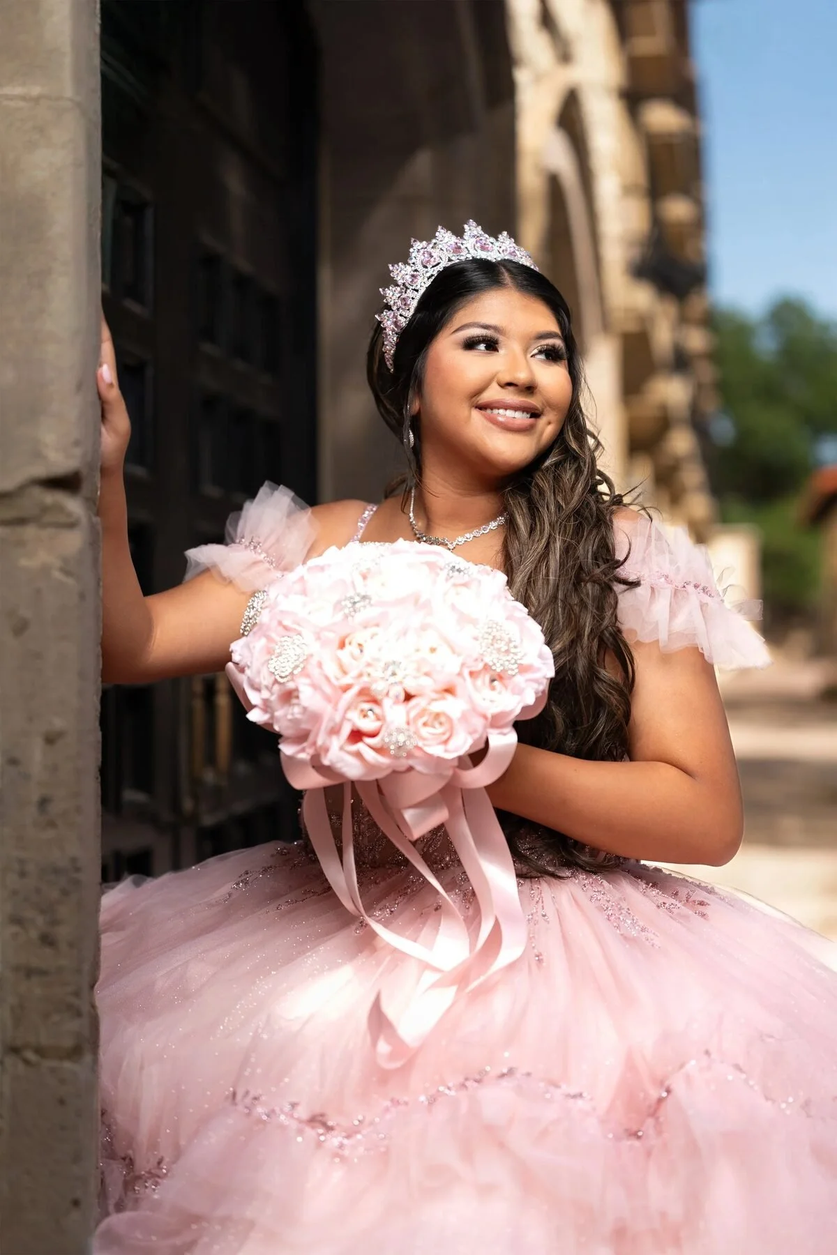 Quinceañera in a blush ball gown holding a rose bouquet in a Dallas doorway