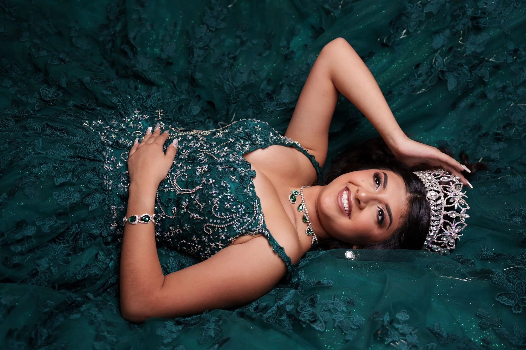 Close editorial portrait of Nevaeh laying on her emerald quinceañera ballgown, showing the beaded bodice detail, emerald necklace, and silver crown