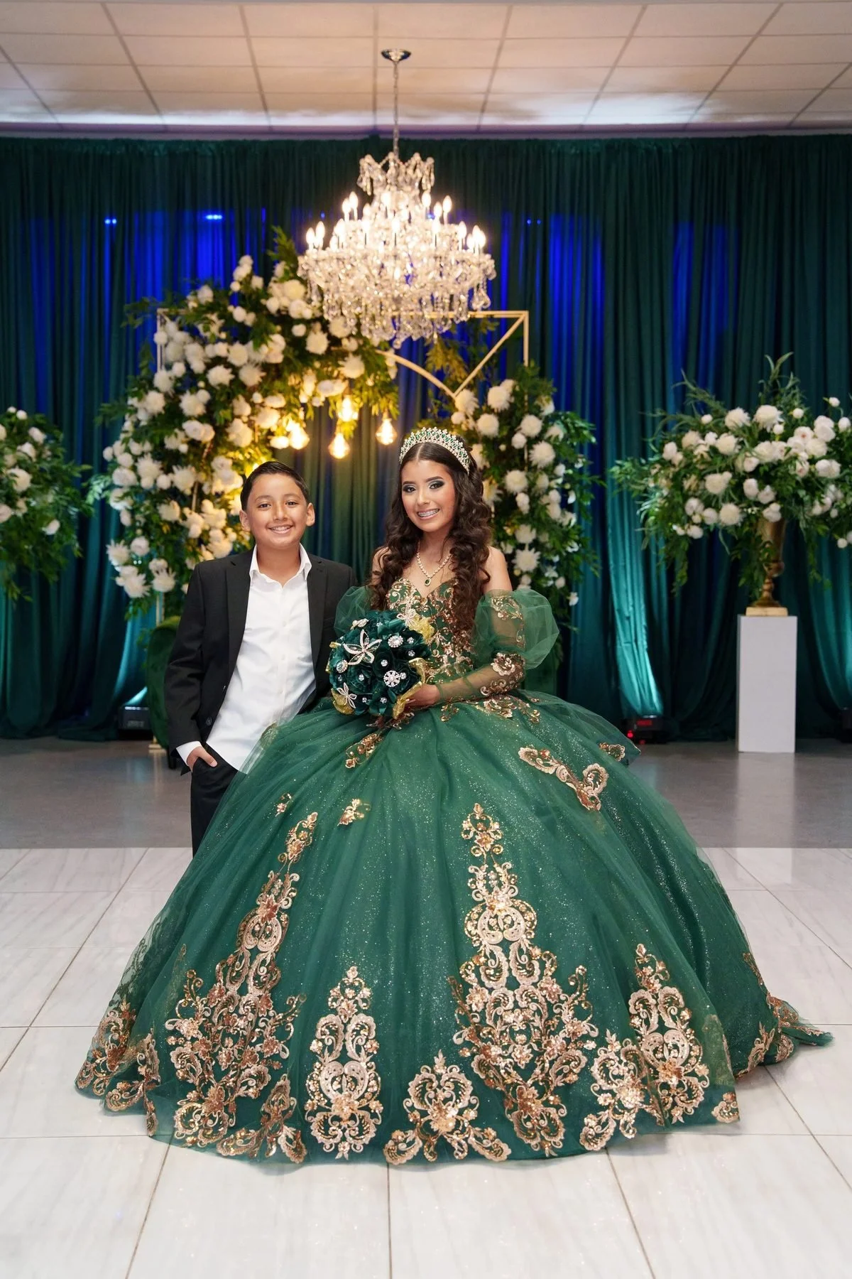 Quinceañera in an emerald and gold ball gown with her chambelán in front of a draped backdrop at a Dallas venue.
