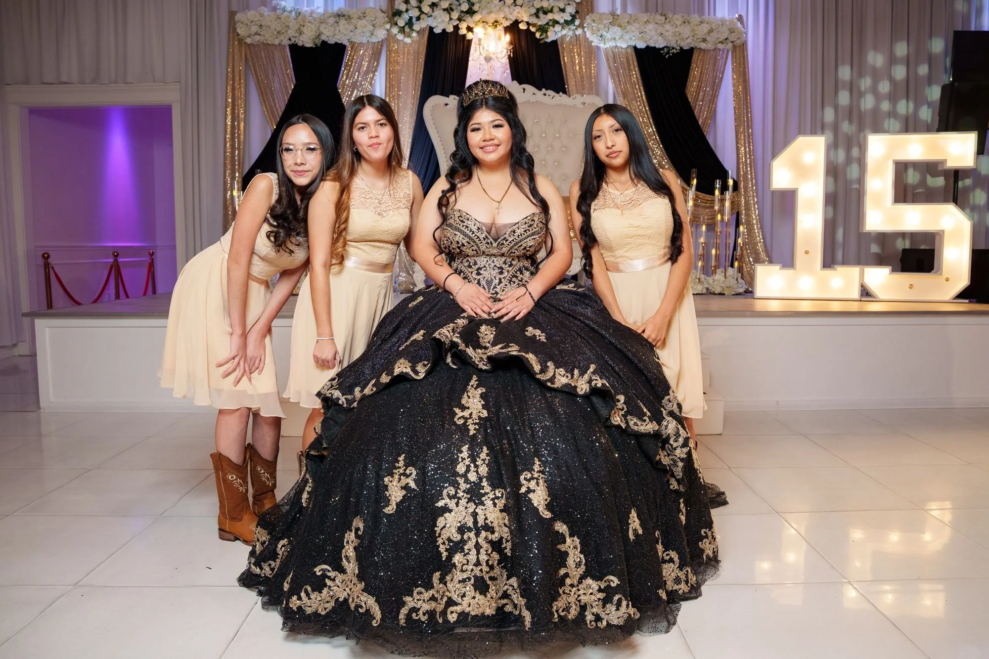 Jayden seated in black and gold ballgown with three damas in cream chiffon and lace dresses, one in cowboy boots, at her quinceañera court portrait at Luxor Banquet Hall Carrollton
