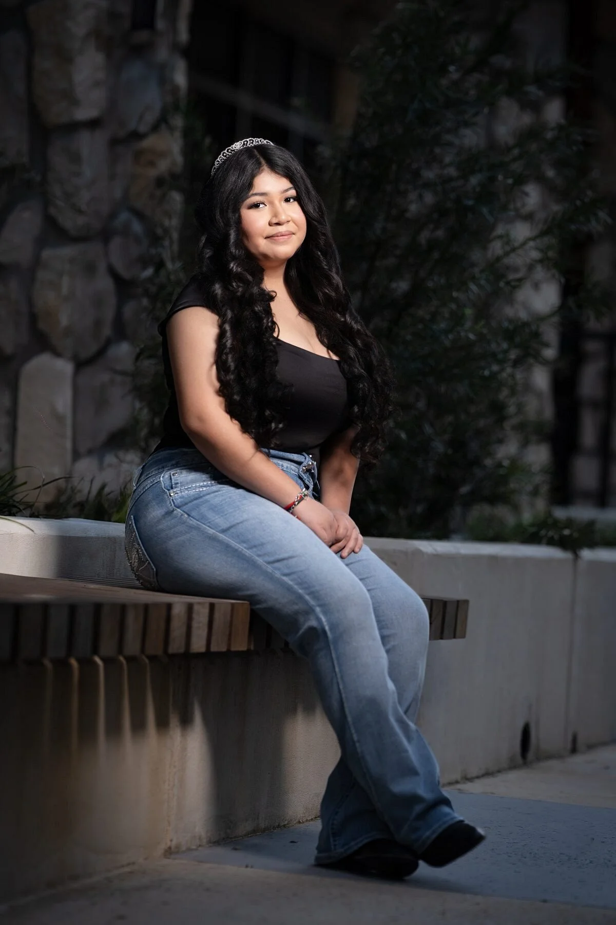 Jayden seated on stone bench in jeans and black top with crystal tiara, casual quinceanera Save the Date portrait in McKinney TX
