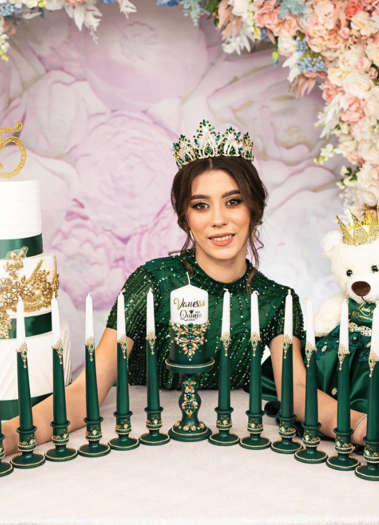 Quinceañera wearing a green gown and crown seated behind a ceremonial candle set with last doll