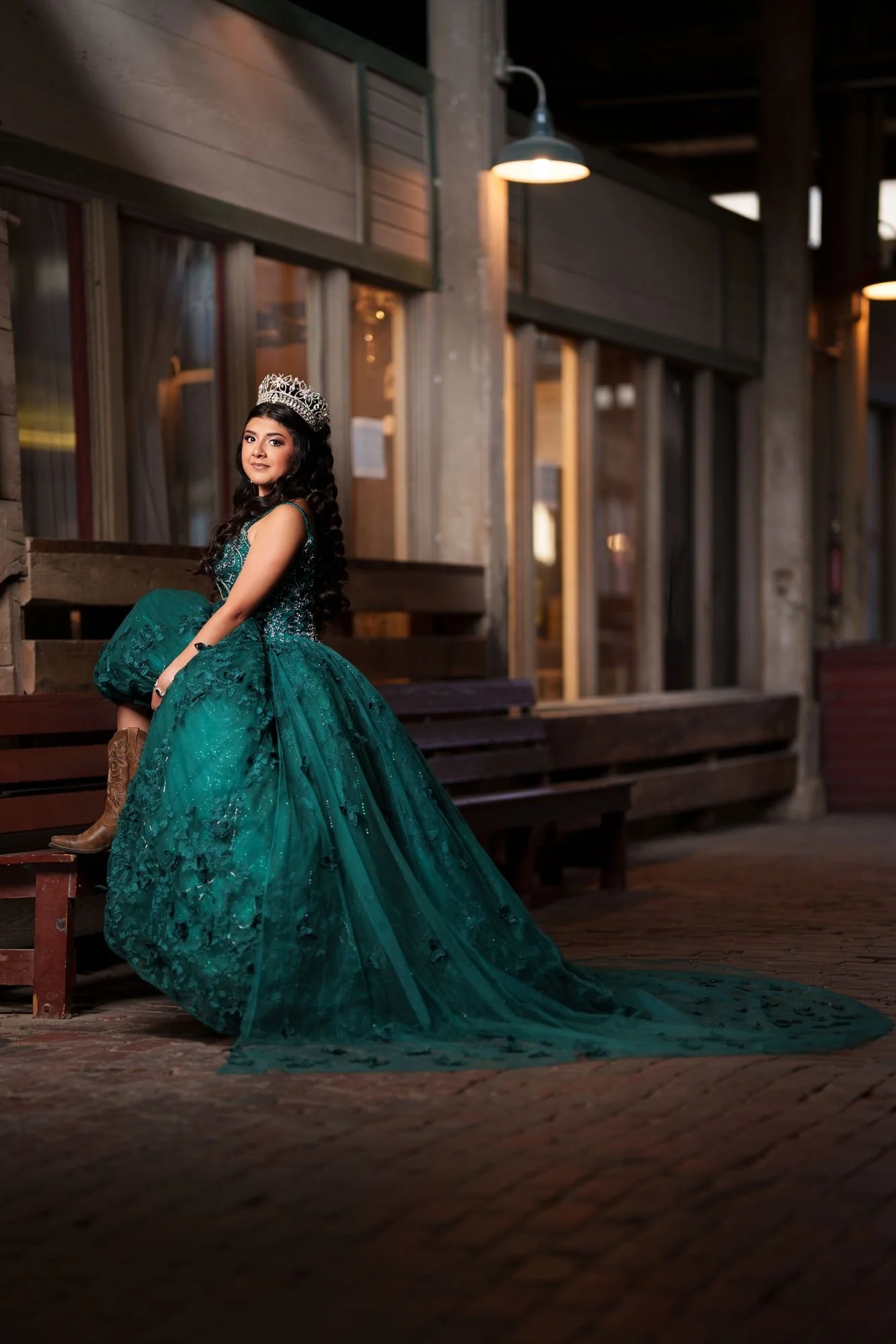 Nevaeh seated on a bench along the historic Stockyards storefronts, emerald ballgown train spread on brick pavement with cowboy boots visible