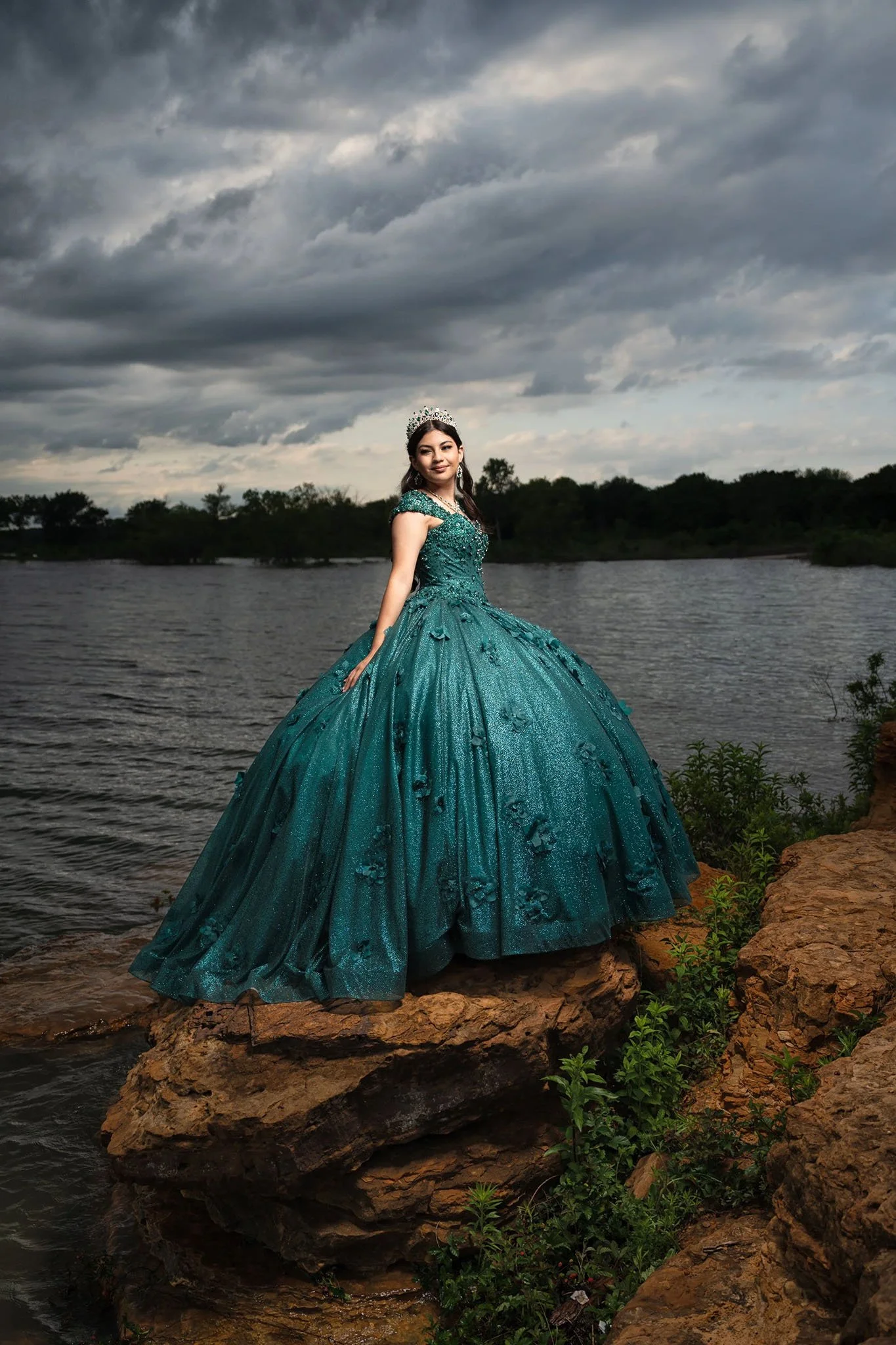 A young woman in a teal ball gown with black floral embellishments, standing on rocks by a body of water, wearing a tiara and earrings, with a cloudy sky overhead.