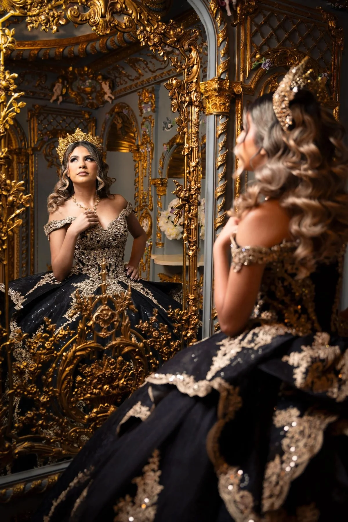 Quinceañera in a black and gold gown admiring her reflection in an ornate gilded mirror at a DFW studio.