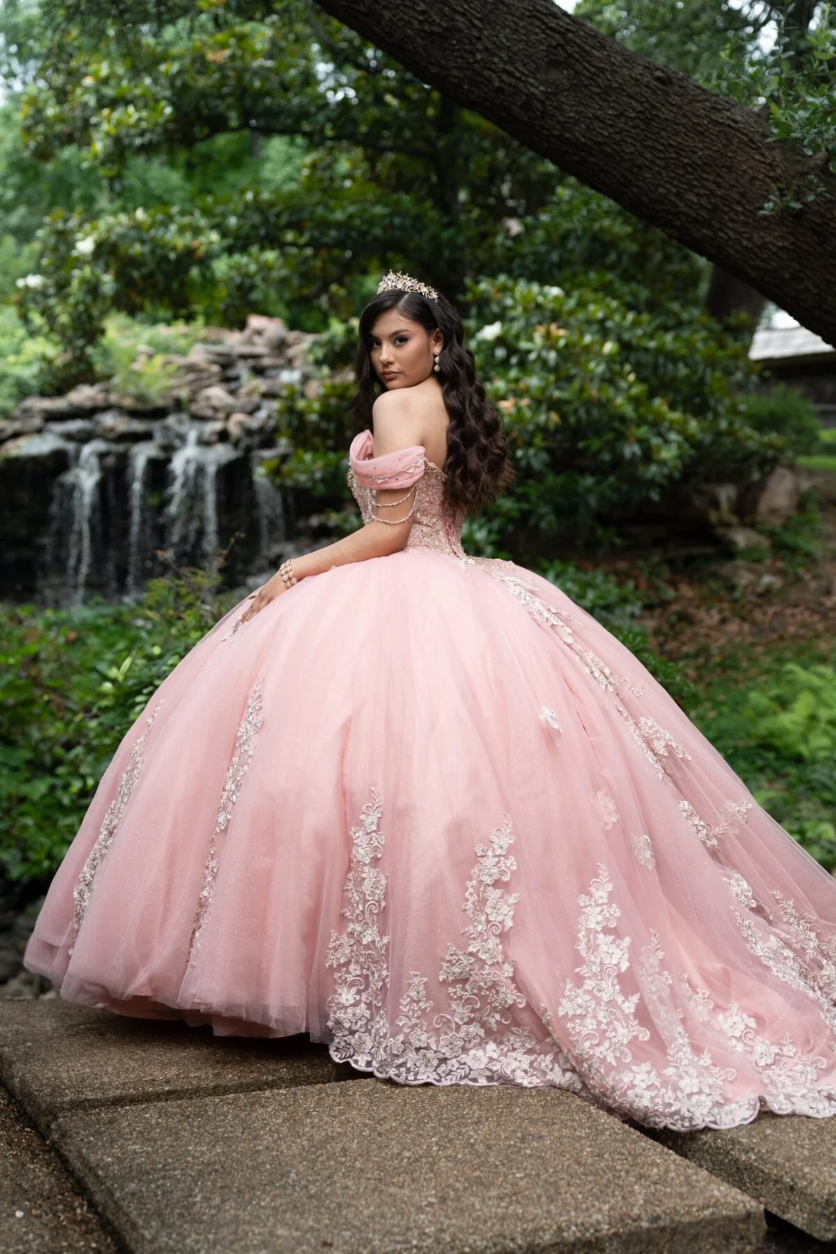A young woman in a pink ball gown with lace details, wearing a tiara and jewelry, standing outdoors near a waterfall and greenery.