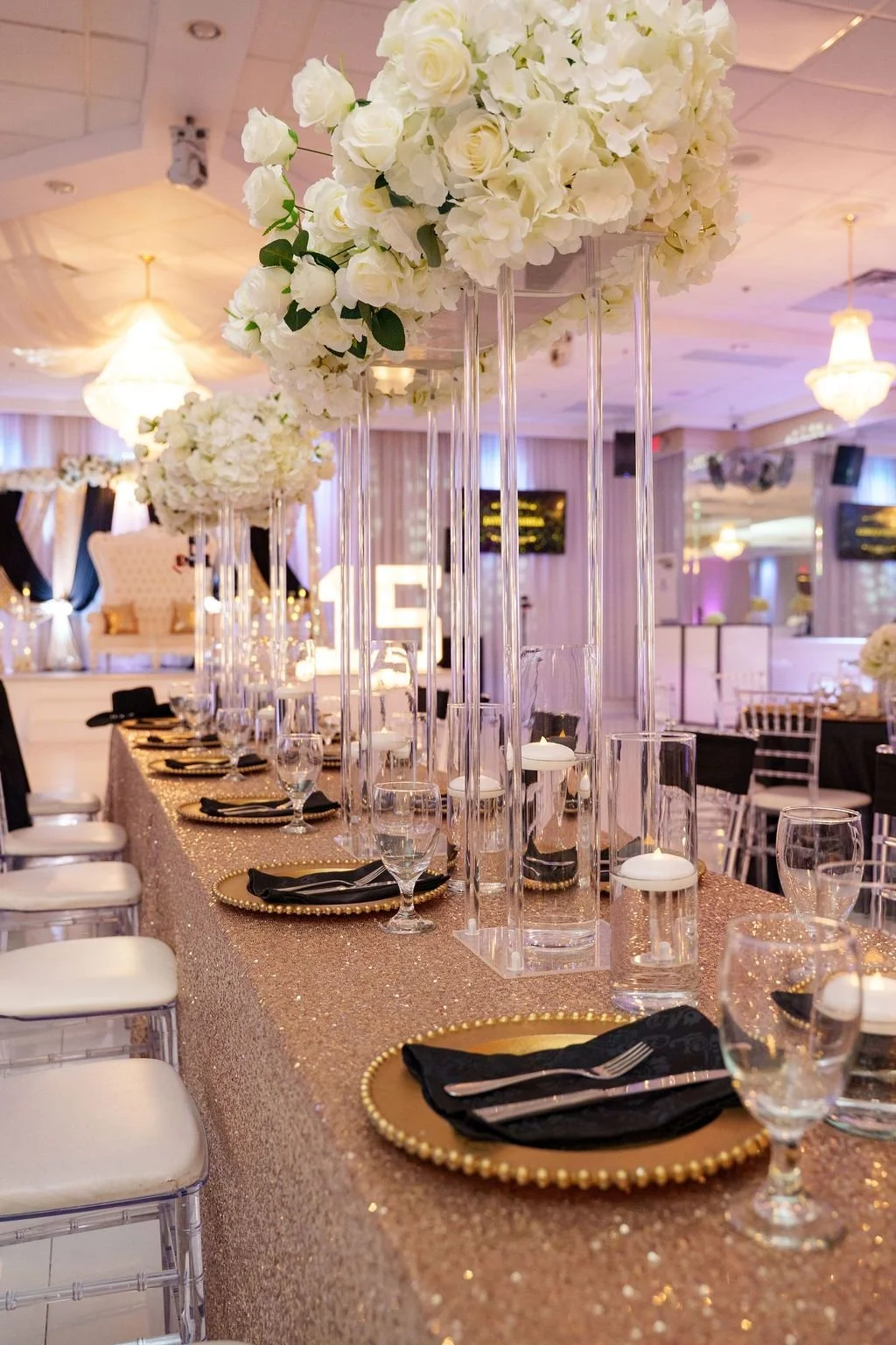 Long head table with rose gold sequin linen tall white floral arrangements and clear chiavari chairs leading toward throne backdrop and marquee 15 letters at Luxor Banquet Hall