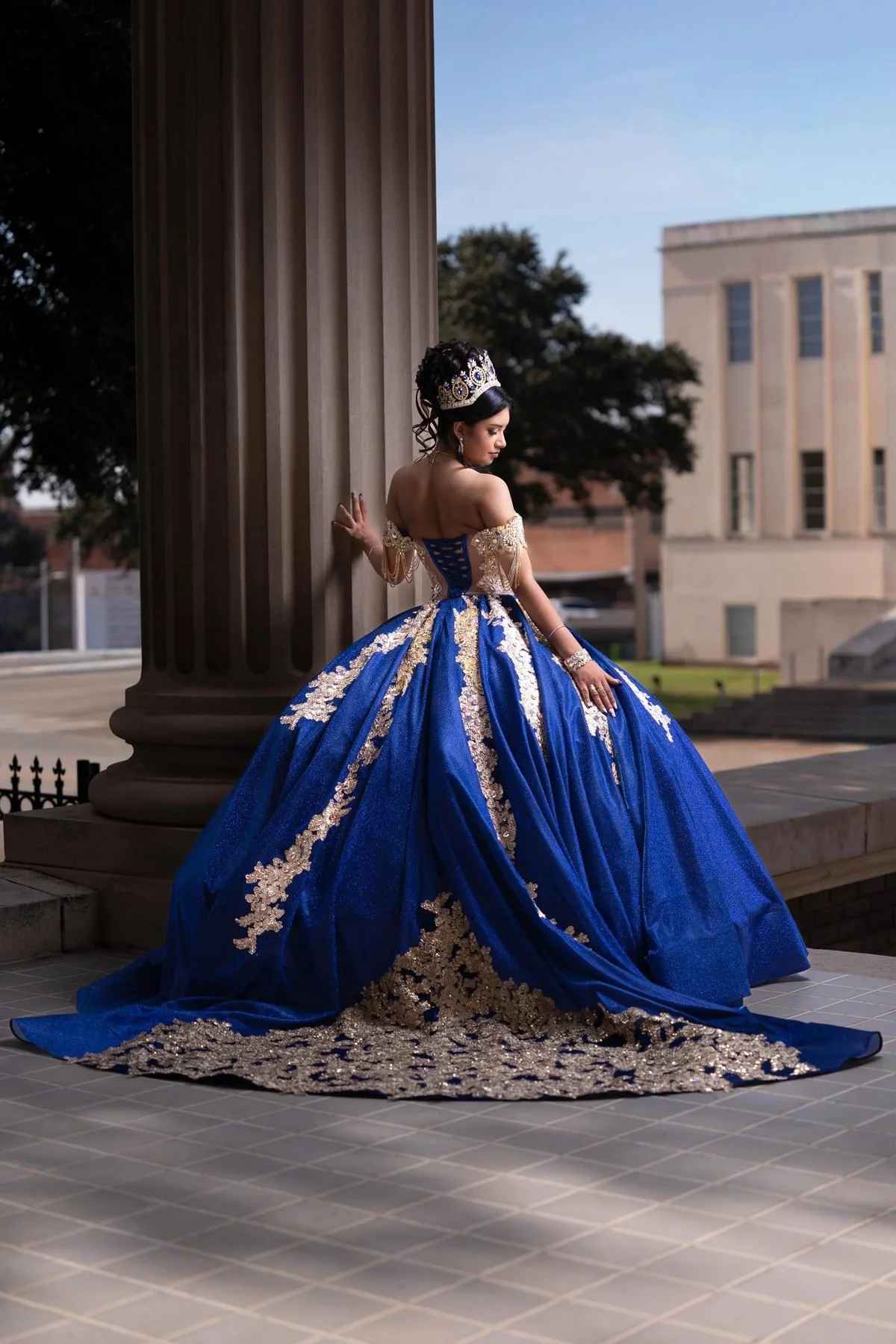 Quinceañera in a royal blue and gold ball gown with a sweeping train beside a courthouse column in DFW.