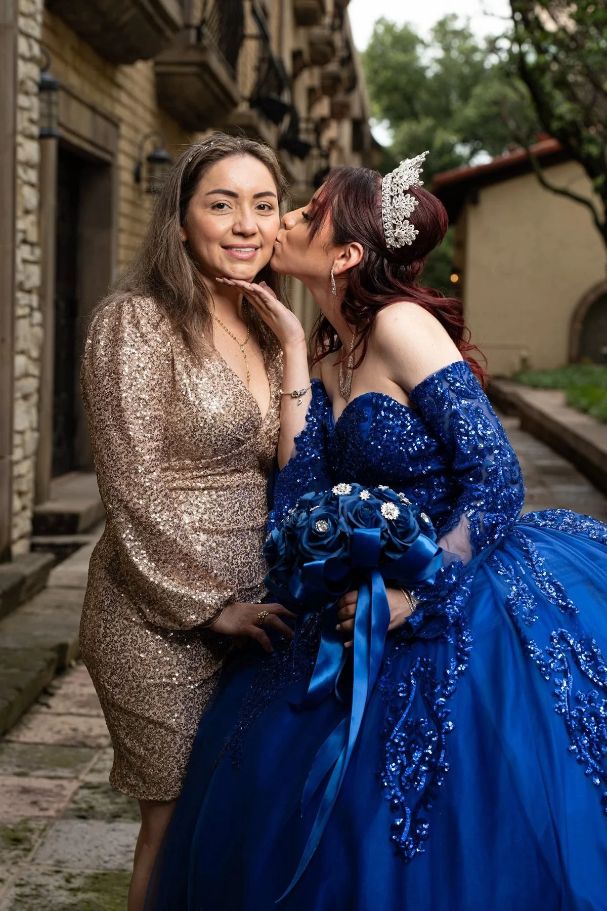 Quinceañera in a royal blue gown kissing her mother on the cheek in a stone courtyard in the DFW area.