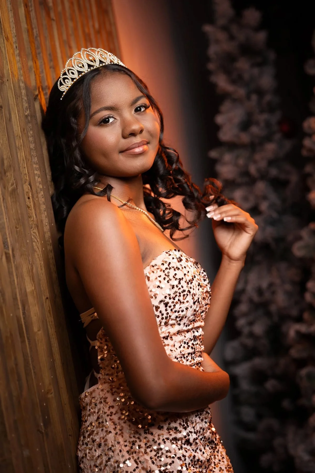 Quinceañera in rose-gold sequin dress and crystal tiara leaning against a wood wall for her DFW save-the-date