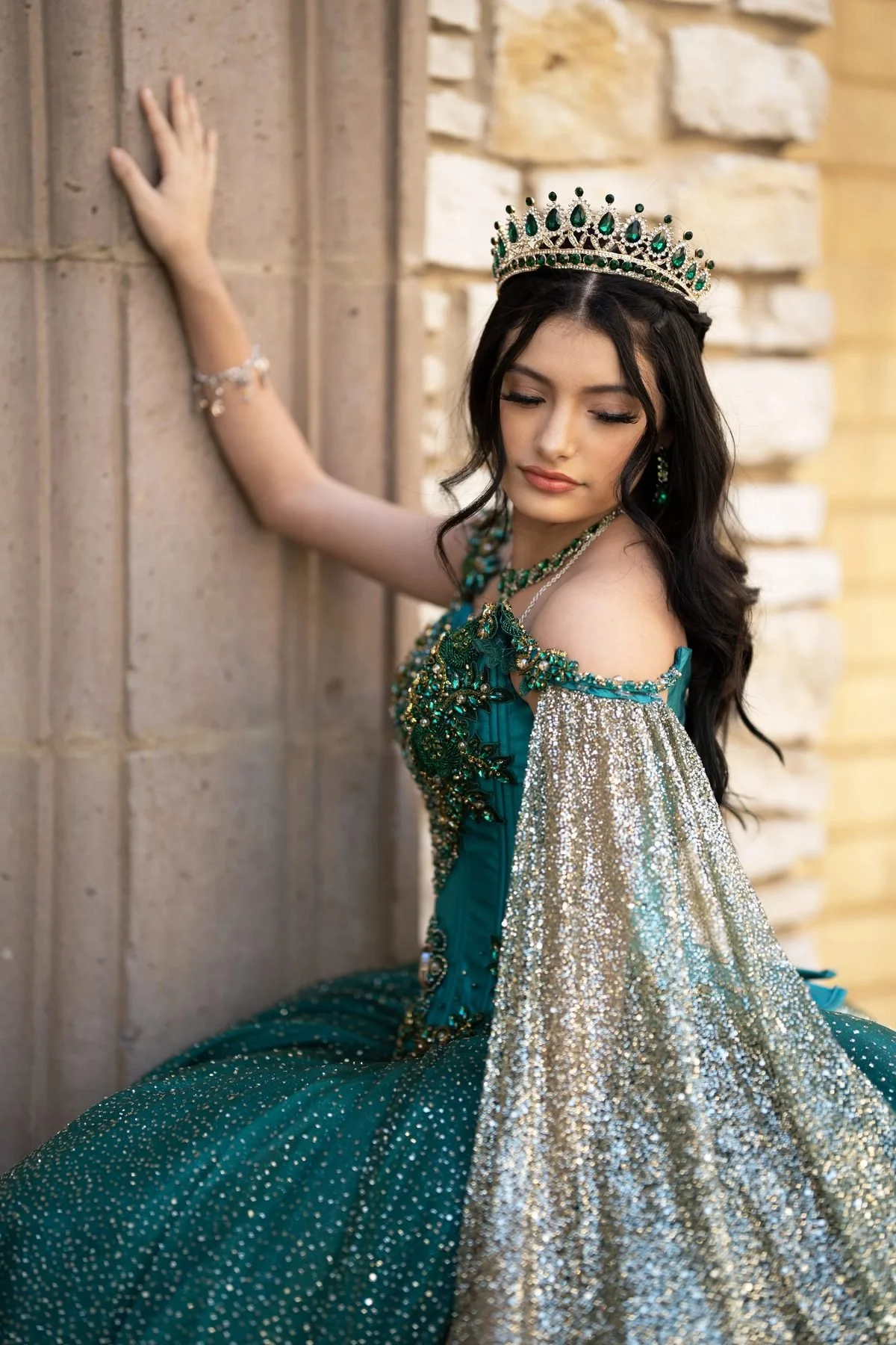 Quinceañera in emerald-green gown with silver and gold sequin cape leaning on a stone wall in DFW