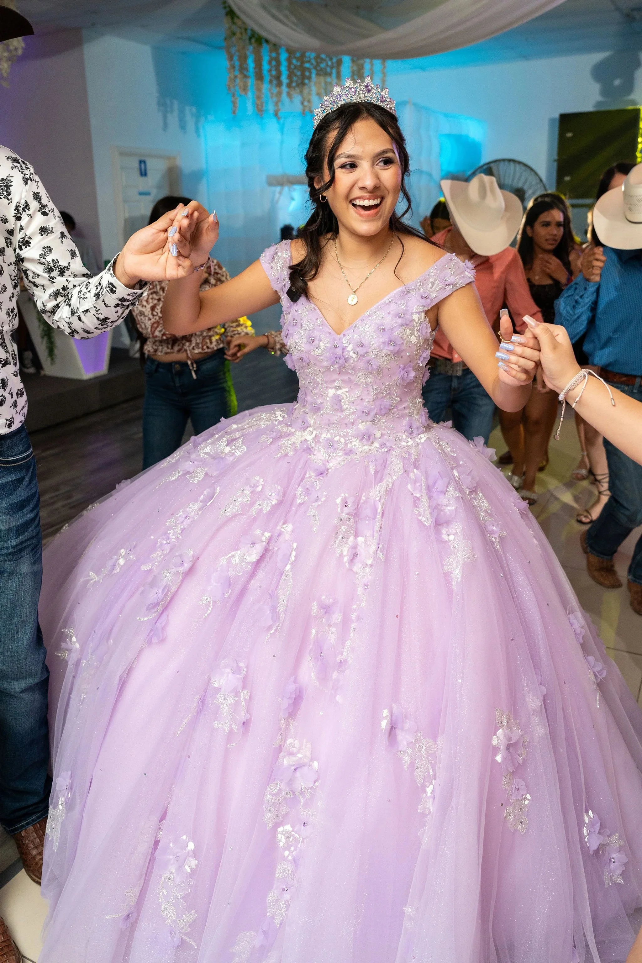 Young woman in a lavender quinceañera dress with floral embroidery, wearing a tiara, dancing and smiling at a celebration.