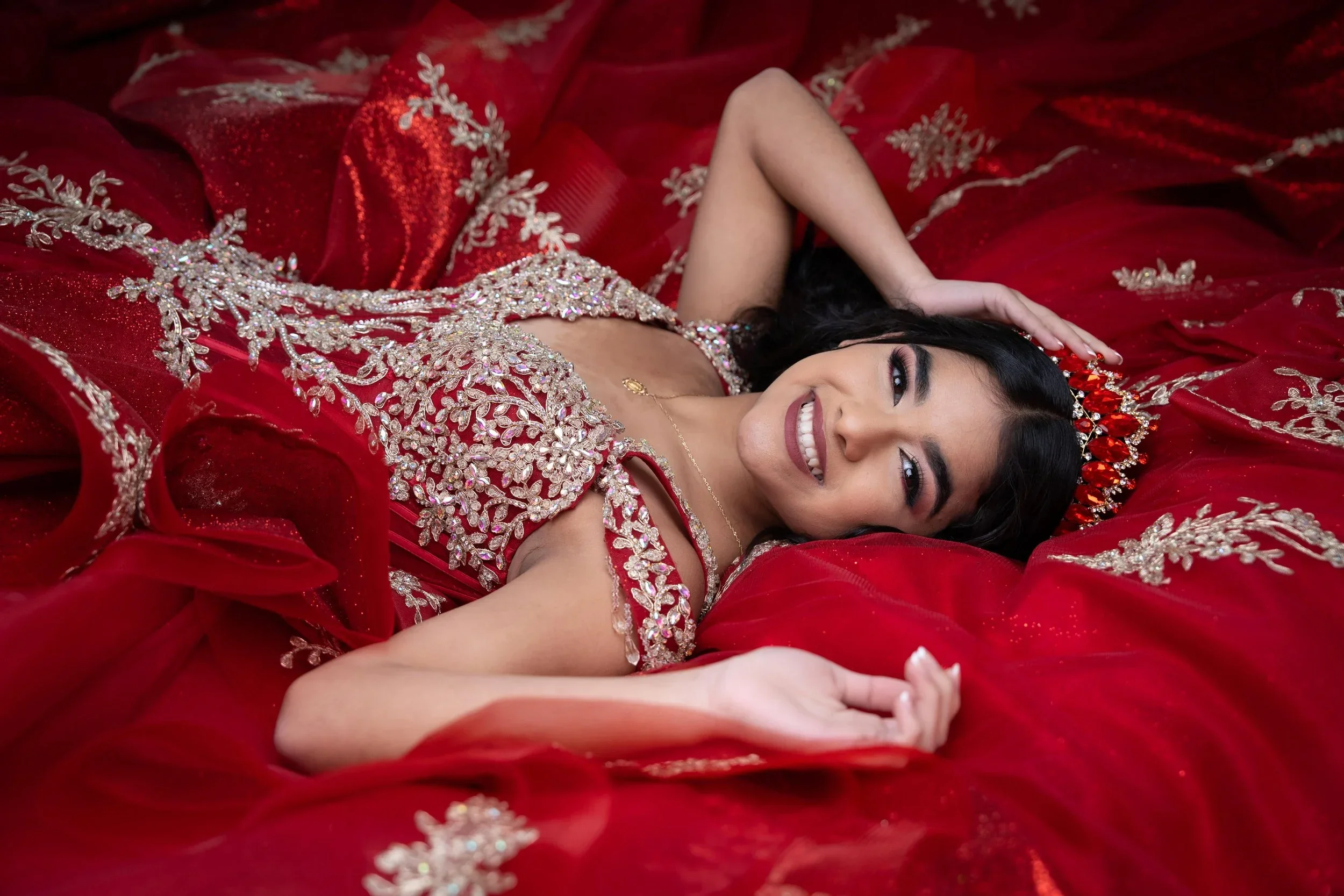 Quinceañera in a red and gold beaded gown reclining among red fabric for a glamorous formal portrait in Dallas–Fort Worth
