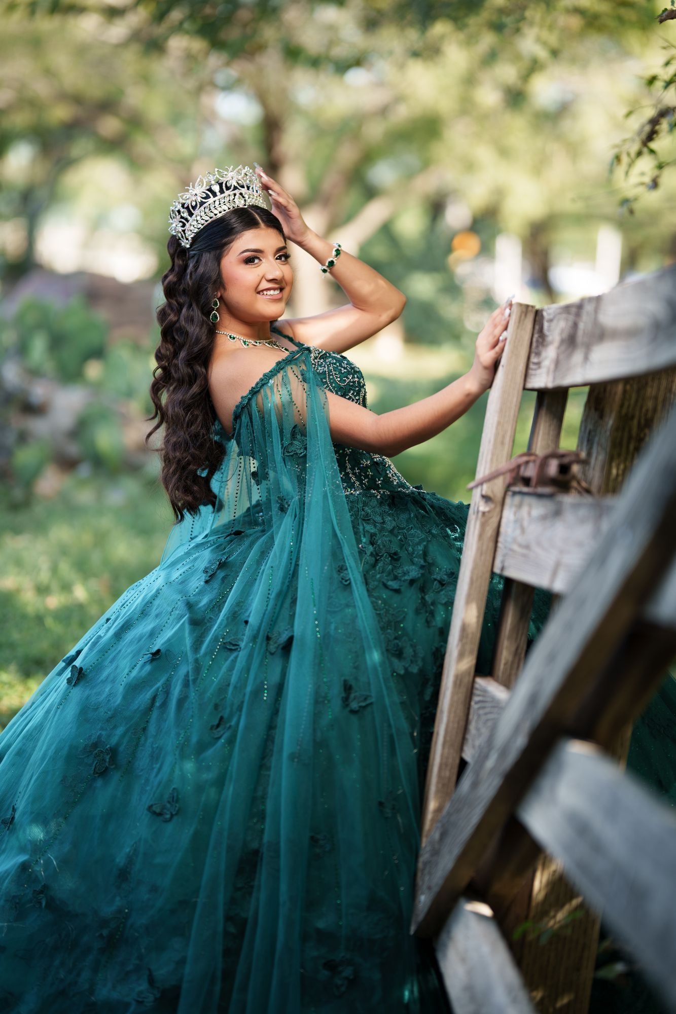 Nevaeh looking back over her shoulder while touching her silver tiara at a wooden gate during her emerald quinceañera preshoot at Fort Worth Stockyards