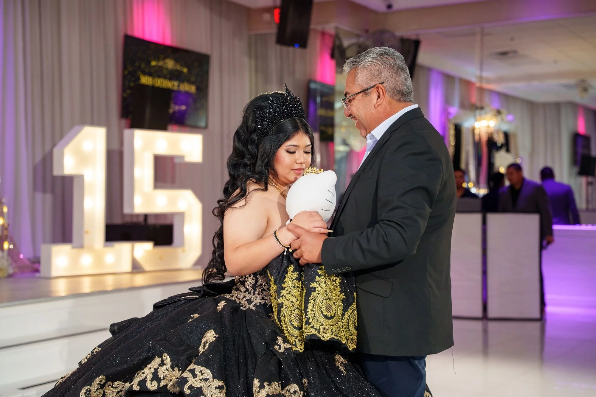 Jayden holding her last doll Hello Kitty plush against her chest during la última muñeca dance with her grandfather in front of marquee 15 letters at her quinceañera at Luxor Banquet Hall Carrollton