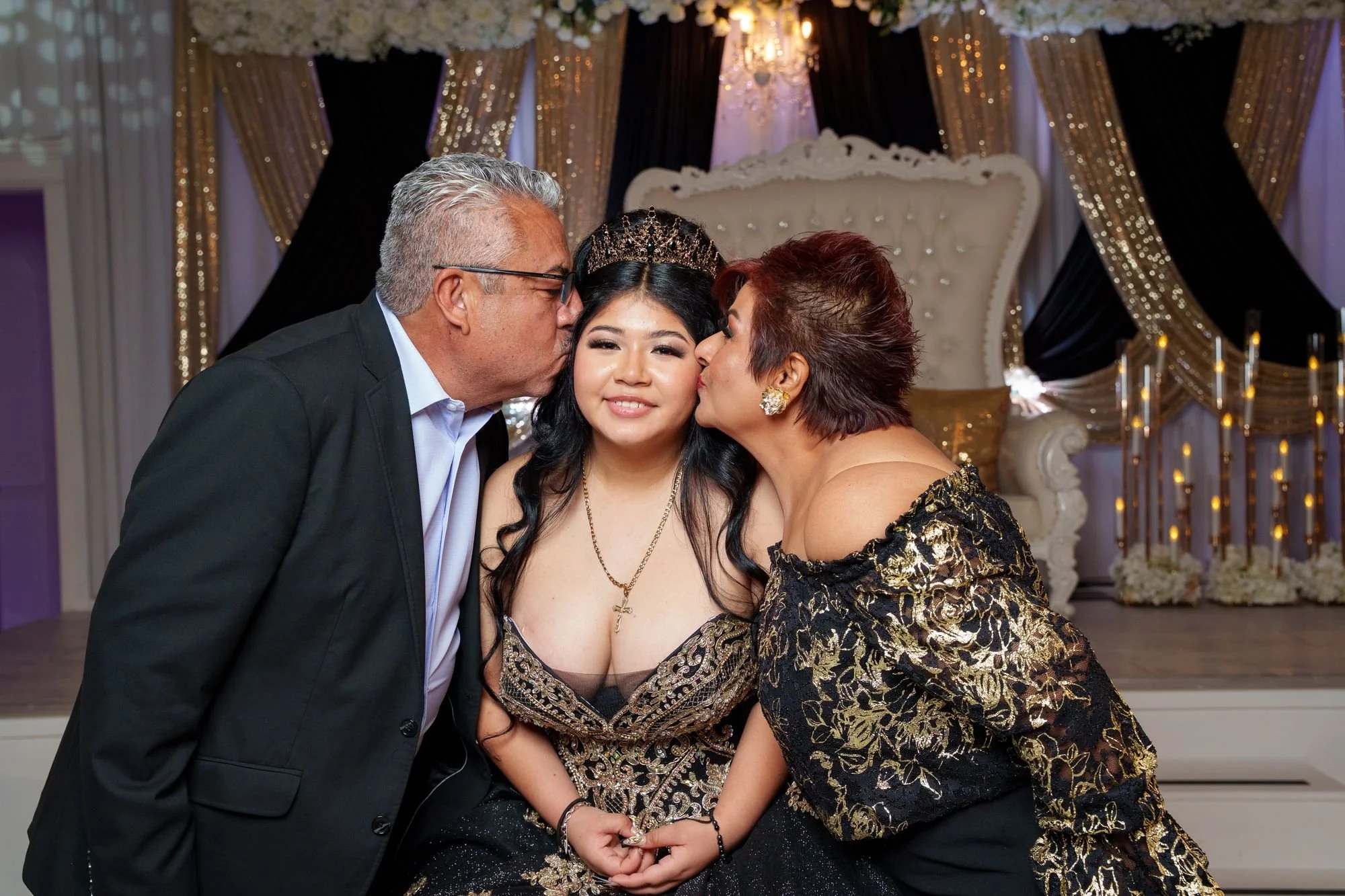 Jayden being kissed on both cheeks by her godparents — silver-haired godfather in dark suit and godmother in black and gold dress — at her quinceañera at Luxor Banquet Hall Carrollton