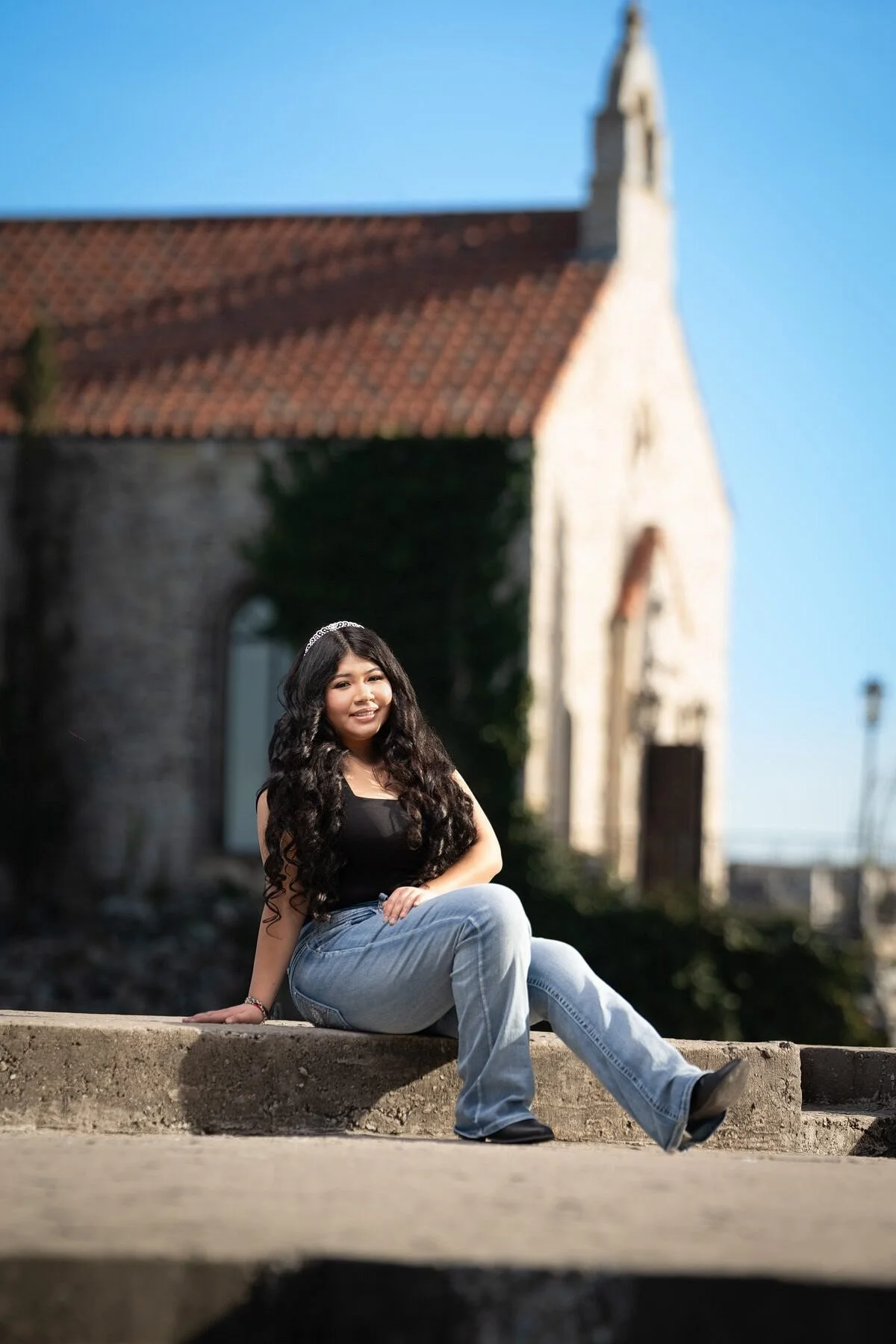 Jayden seated on stone steps in jeans and tank top, Mediterranean chapel and tile roof behind her at Adriatica Village McKinney