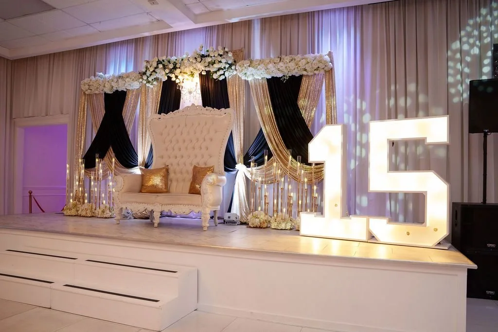 White tufted throne with gold pillows on stage under black and gold draped backdrop with white floral canopy beside marquee 15 letters at Jayden's quinceañera at Luxor Banquet Hall Carrollton