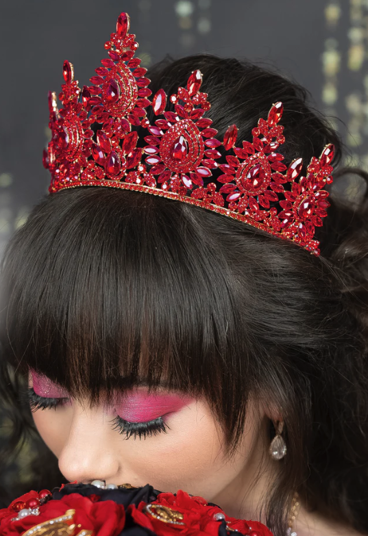Close-up of a young woman wearing an ornate red jeweled crown, with dark hair styled in an updo and bold pink eye makeup, holding a bouquet of red roses.