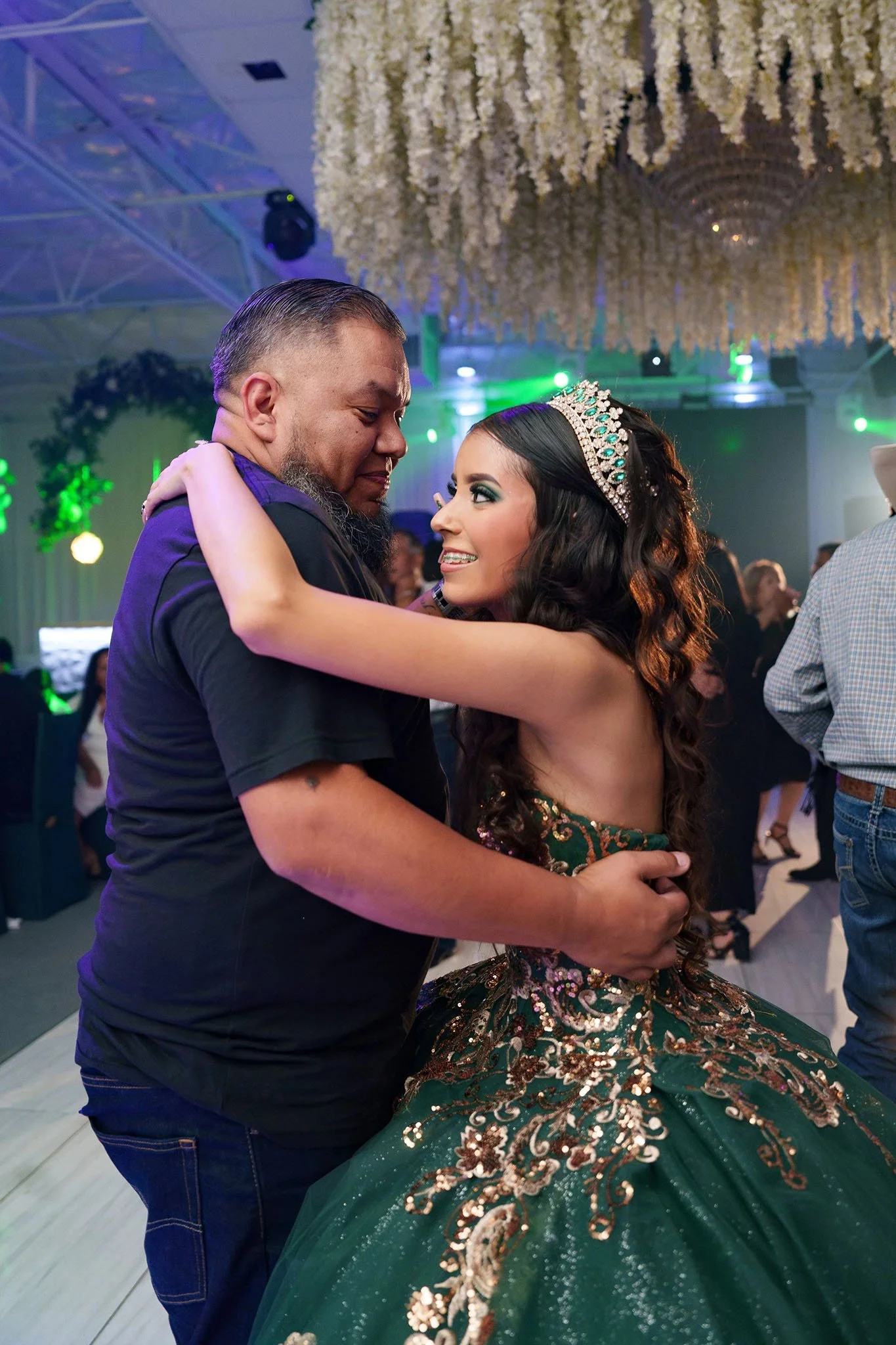 A young woman in a green and gold embellished ball gown with a tiara, dancing with an older man at a celebration or dance event.
