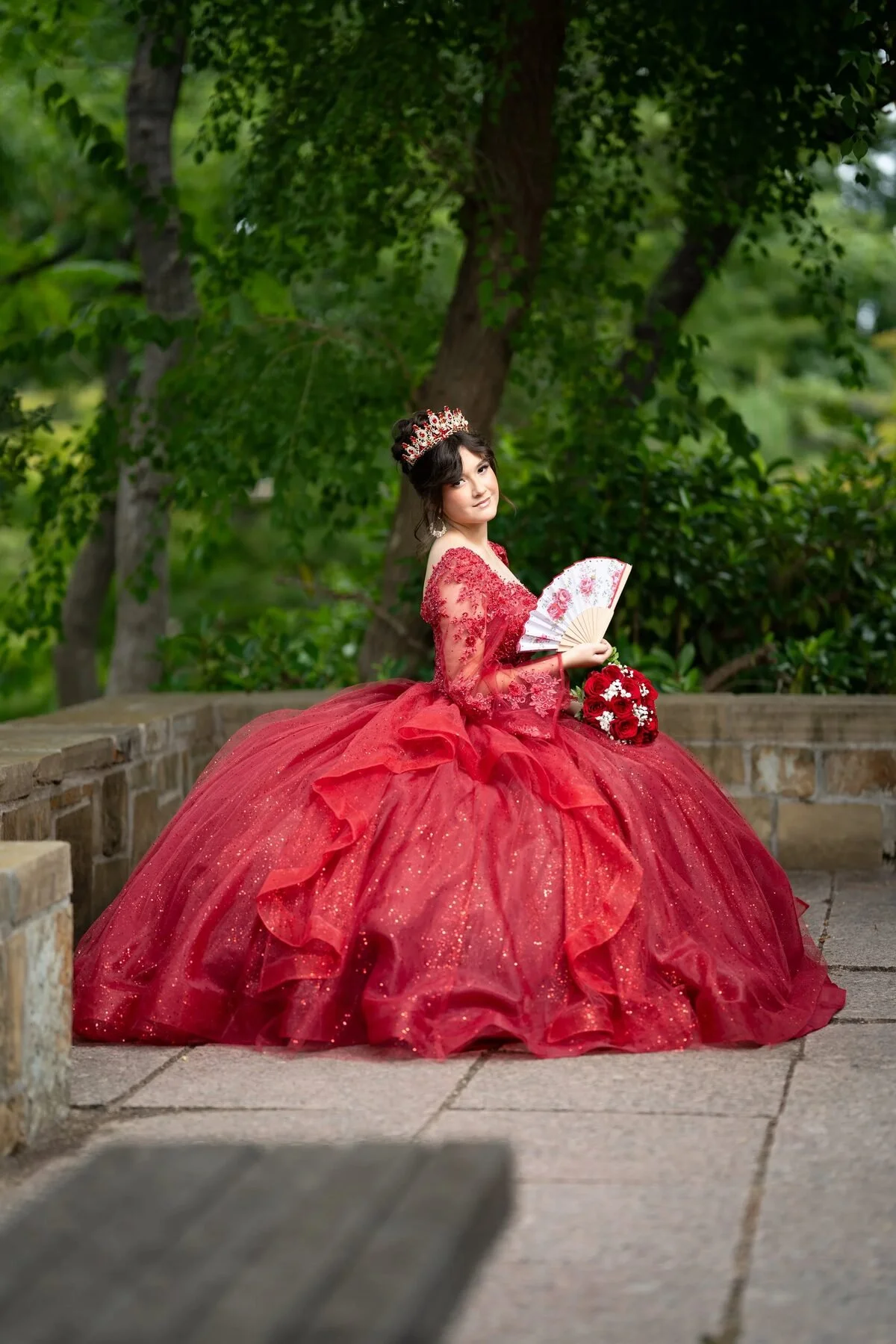 Quinceañera in a red ball gown and crown holding a fan in a Dallas garden