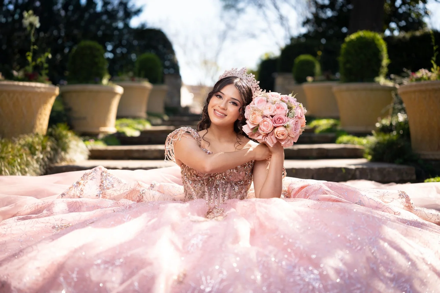 Preshoot formal de quinceañera con vestido rosa y tiara de cristal en un jardín formal del DFW, retrato estilo princesa con ramo de rosas rosas por New Dawn Photo