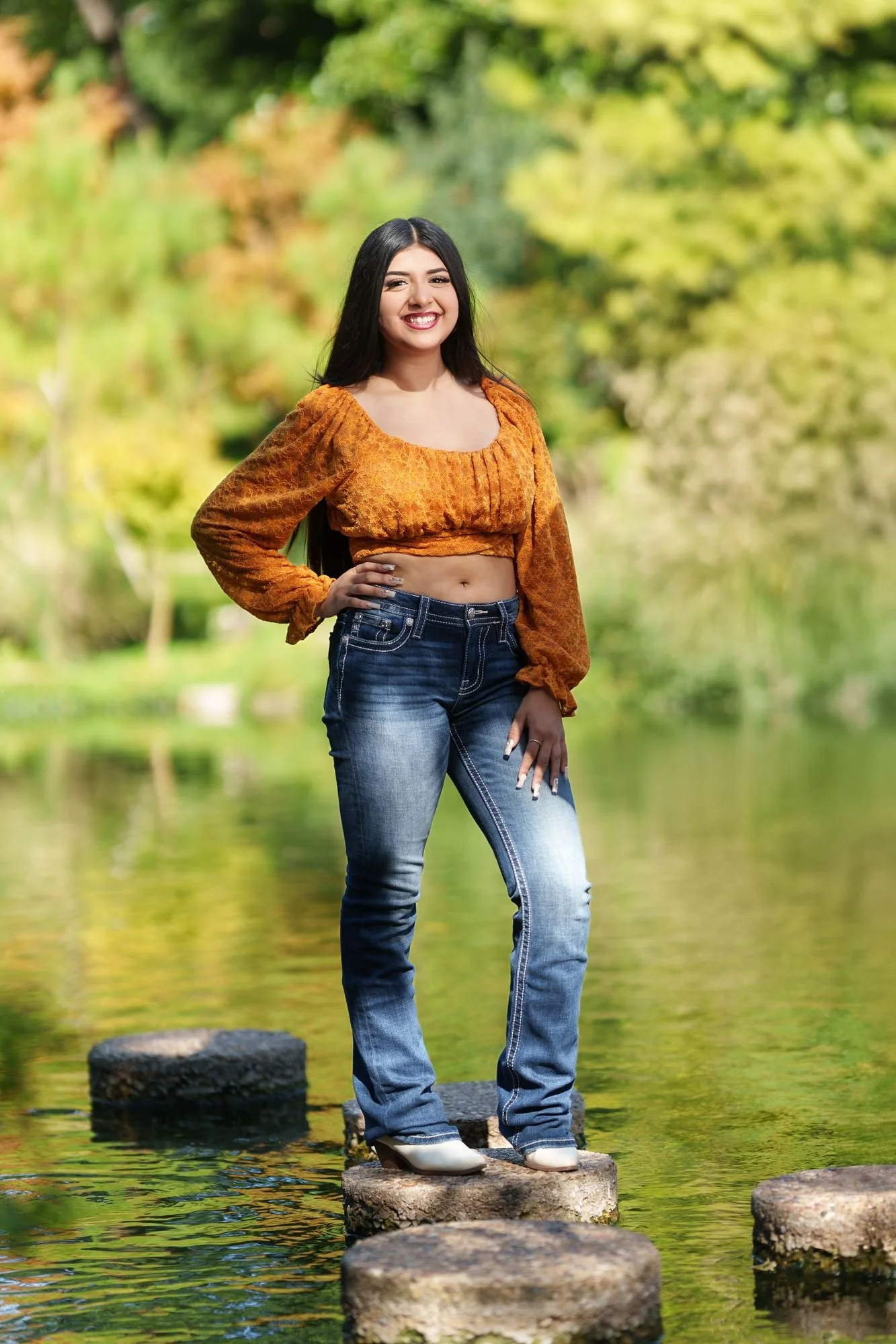 Nevaeh standing on stepping stones across the koi pond at Fort Worth Botanic Garden in a burnt orange boho western Save the Date quinceañera outfit