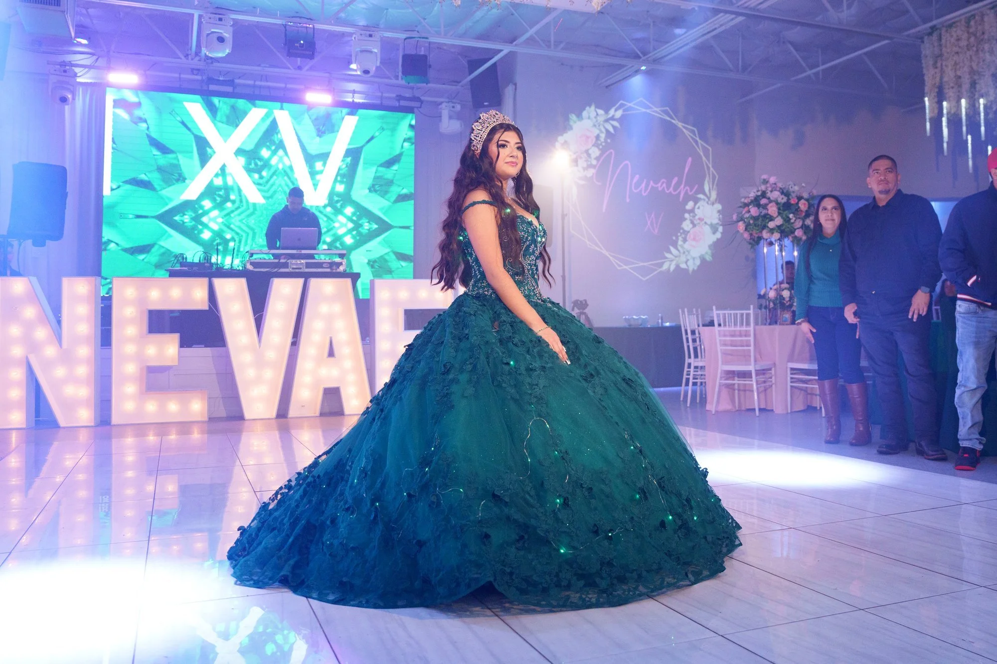 Nevaeh standing alone on the dance floor in her emerald ballgown and crown, surrounded by her illuminated NEVAEH marquee letters, custom XV screen, and floral monogram — the quinceañera owning her moment