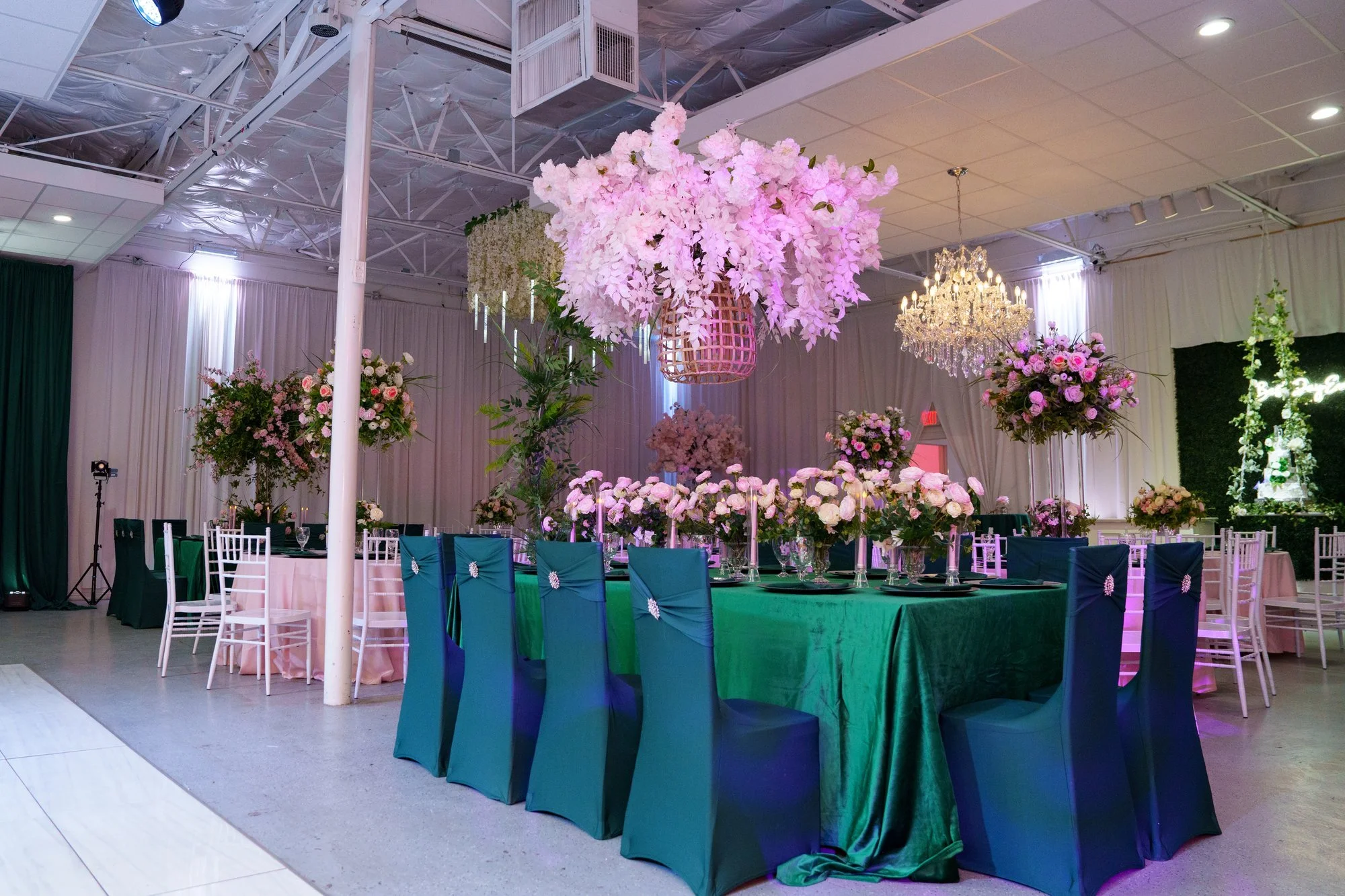 Wide view of Nevaeh's quinceañera reception at Blanc Event Center in Dallas — emerald velvet banquet table, pink rose centerpieces, hanging pink floral chandelier, and crystal chandelier overhead
