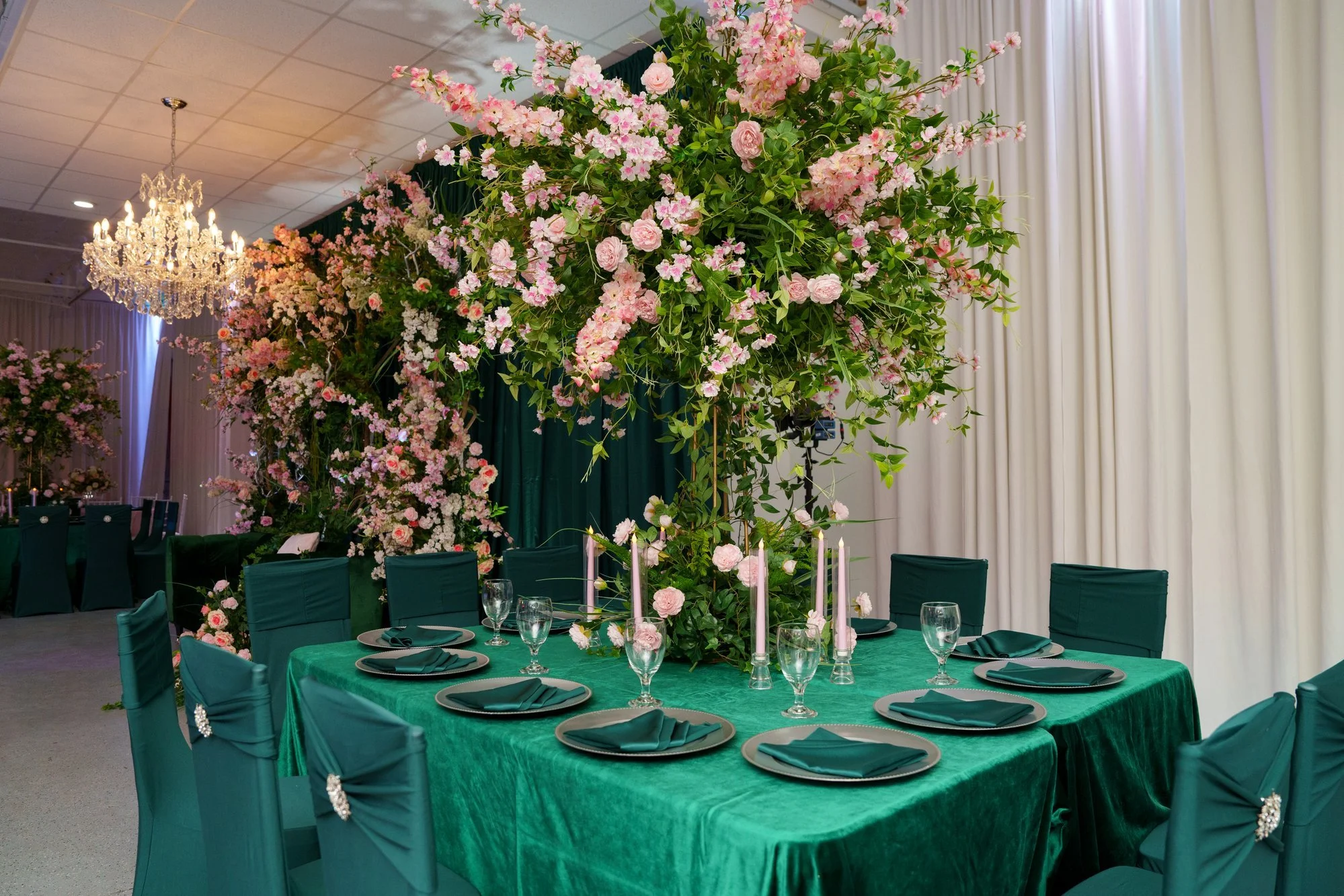 Long banquet table with emerald green runners, crystal chandeliers, and pink floral centerpieces at Nevaeh's quinceañera reception