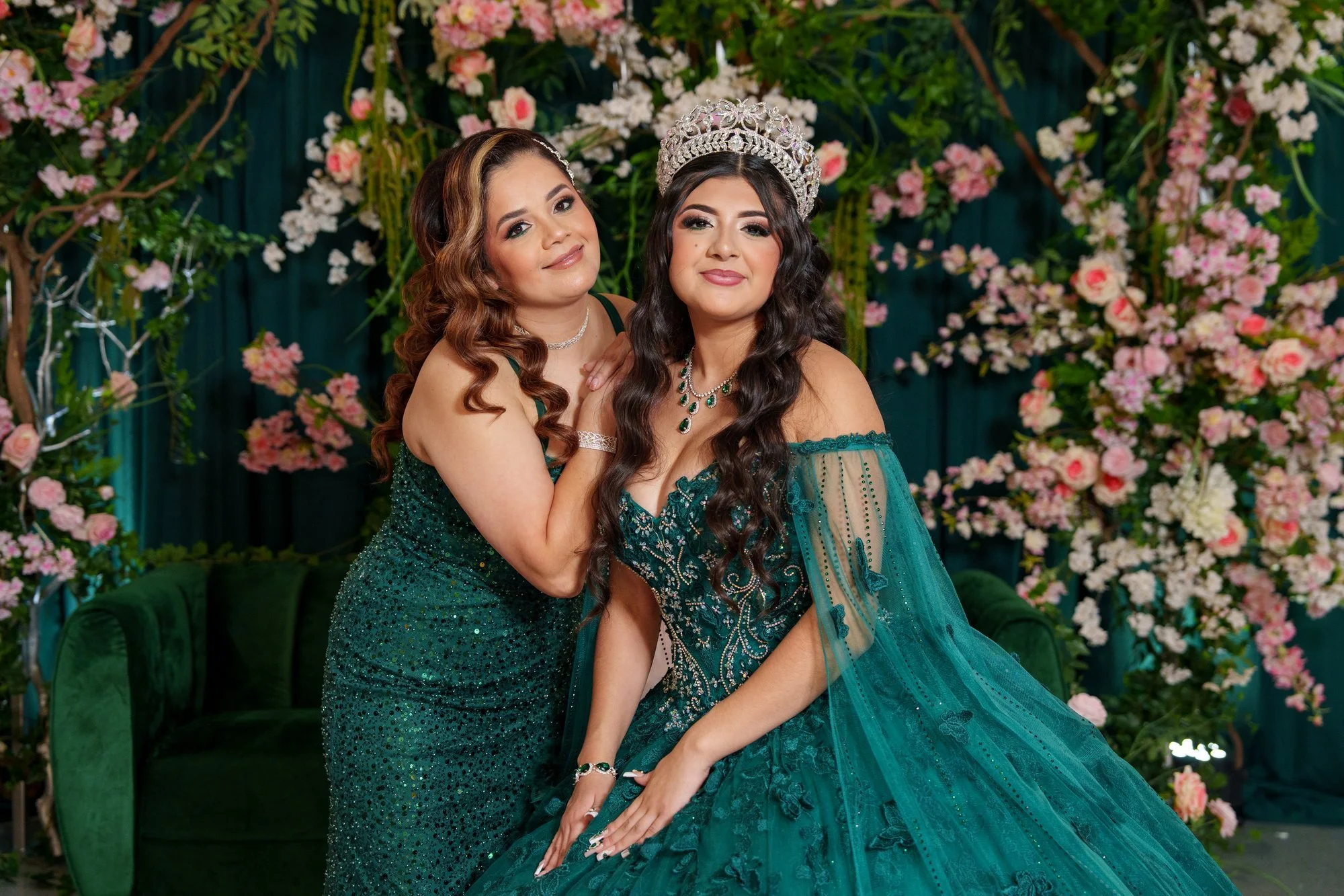 Nevaeh and her mother in front of a pink floral arch with emerald velvet backdrop, Nevaeh wearing her crown and ballgown, mother wearing emerald sequined dress
