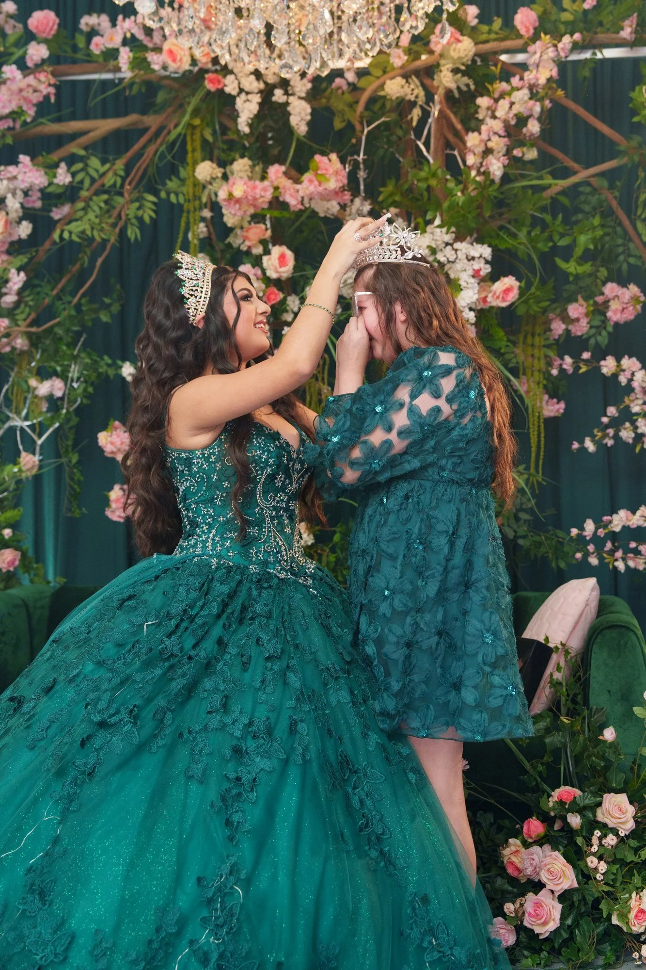 Nevaeh placing a crown on her younger sister's head during the traditional passing-forward moment, pink florals behind them, her sister overcome with emotion