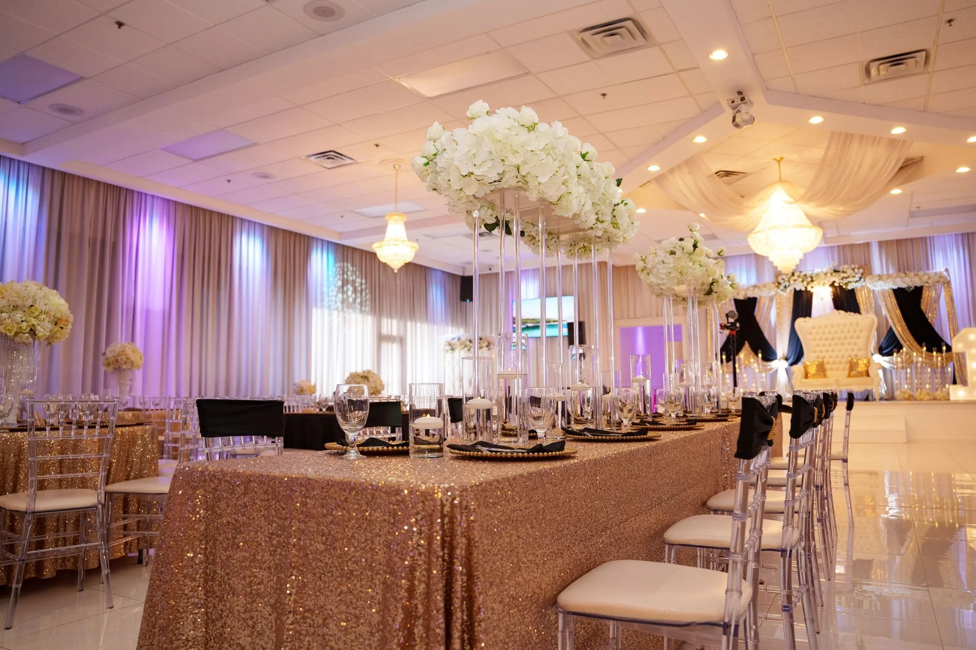 Long head table set with rose gold sequins and white floral arrangements leading to throne under black and gold draped backdrop with white rose canopy at Luxor Banquet Hall