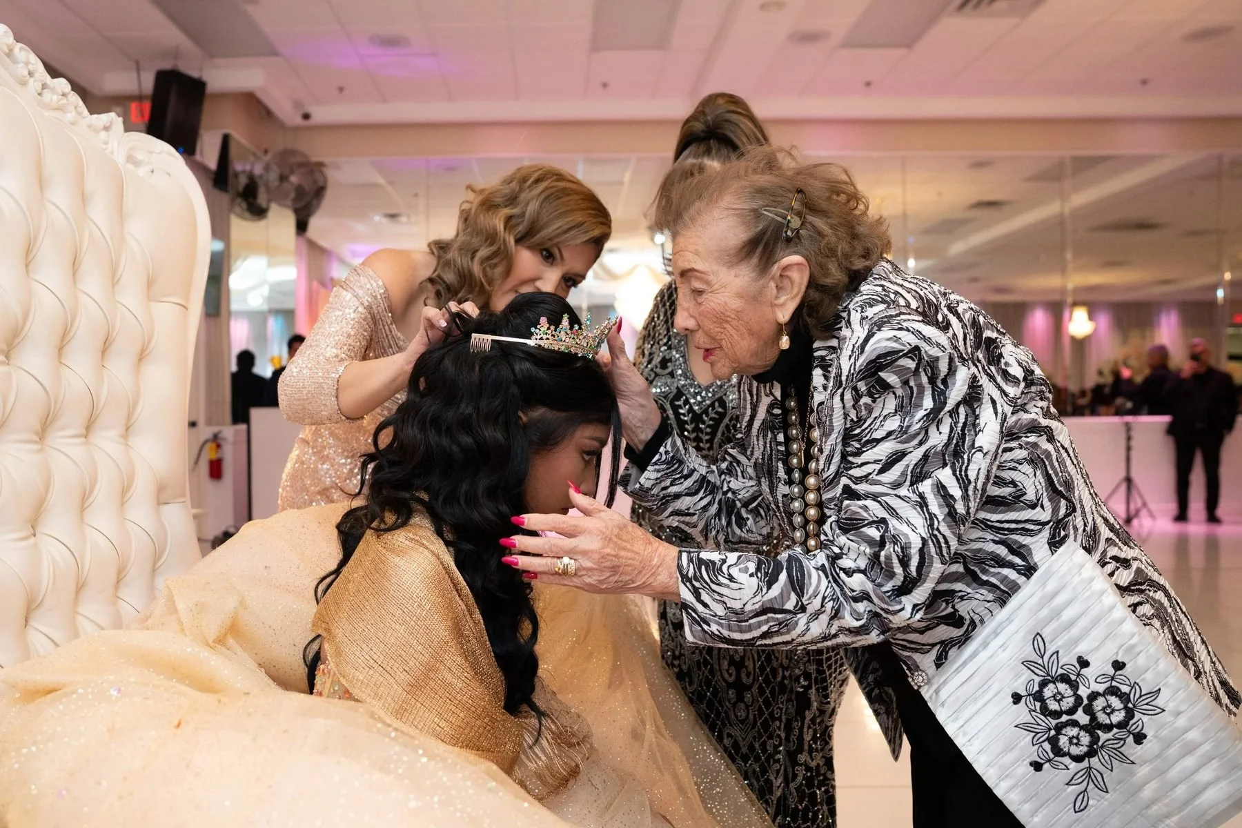 Quinceañera kneeling in her champagne gown as her mother and grandmother place the crown during the DFW crowning ceremony