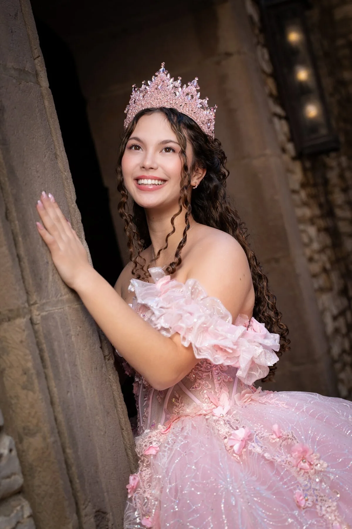 Quinceanera in a pink gown and crown poses by a stone archway in Dallas.