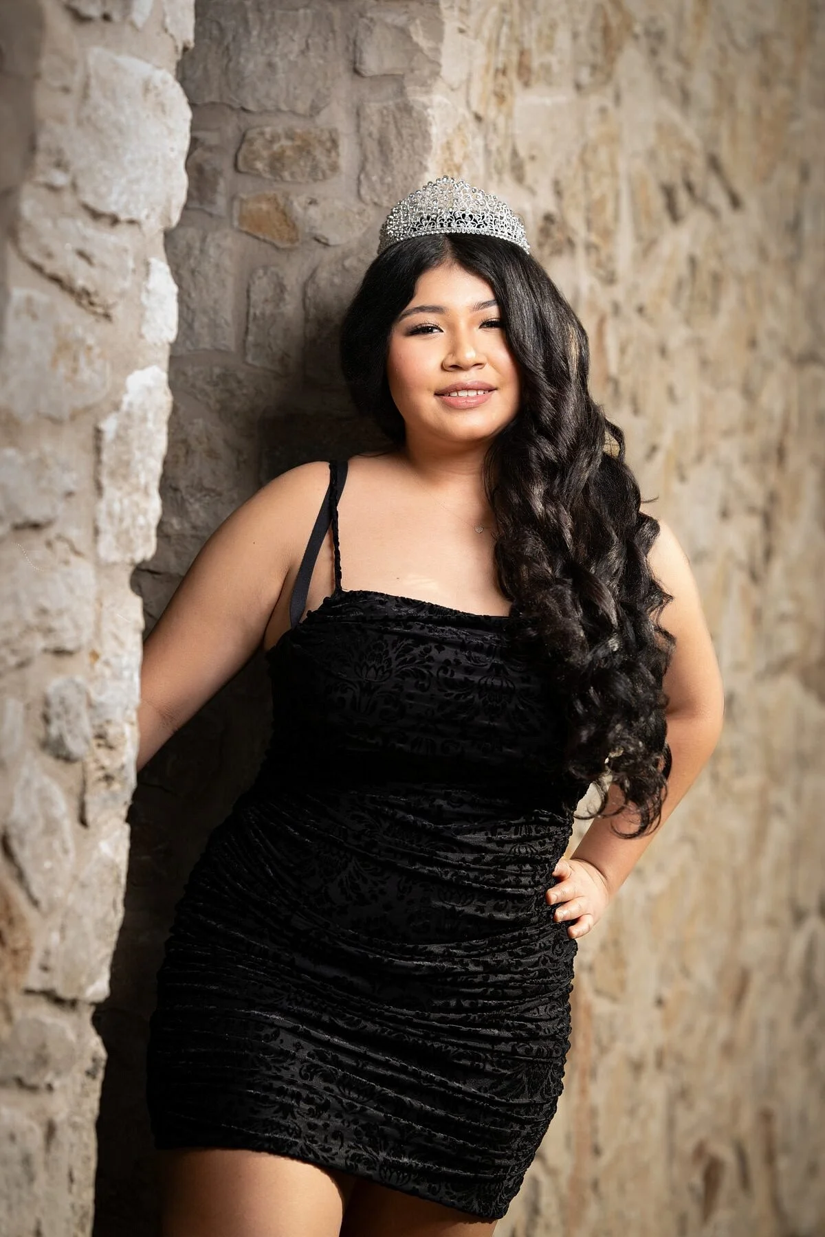 Jayden in black velvet quinceanera dress and crystal tiara leaning against limestone wall at Adriatica Village McKinney
