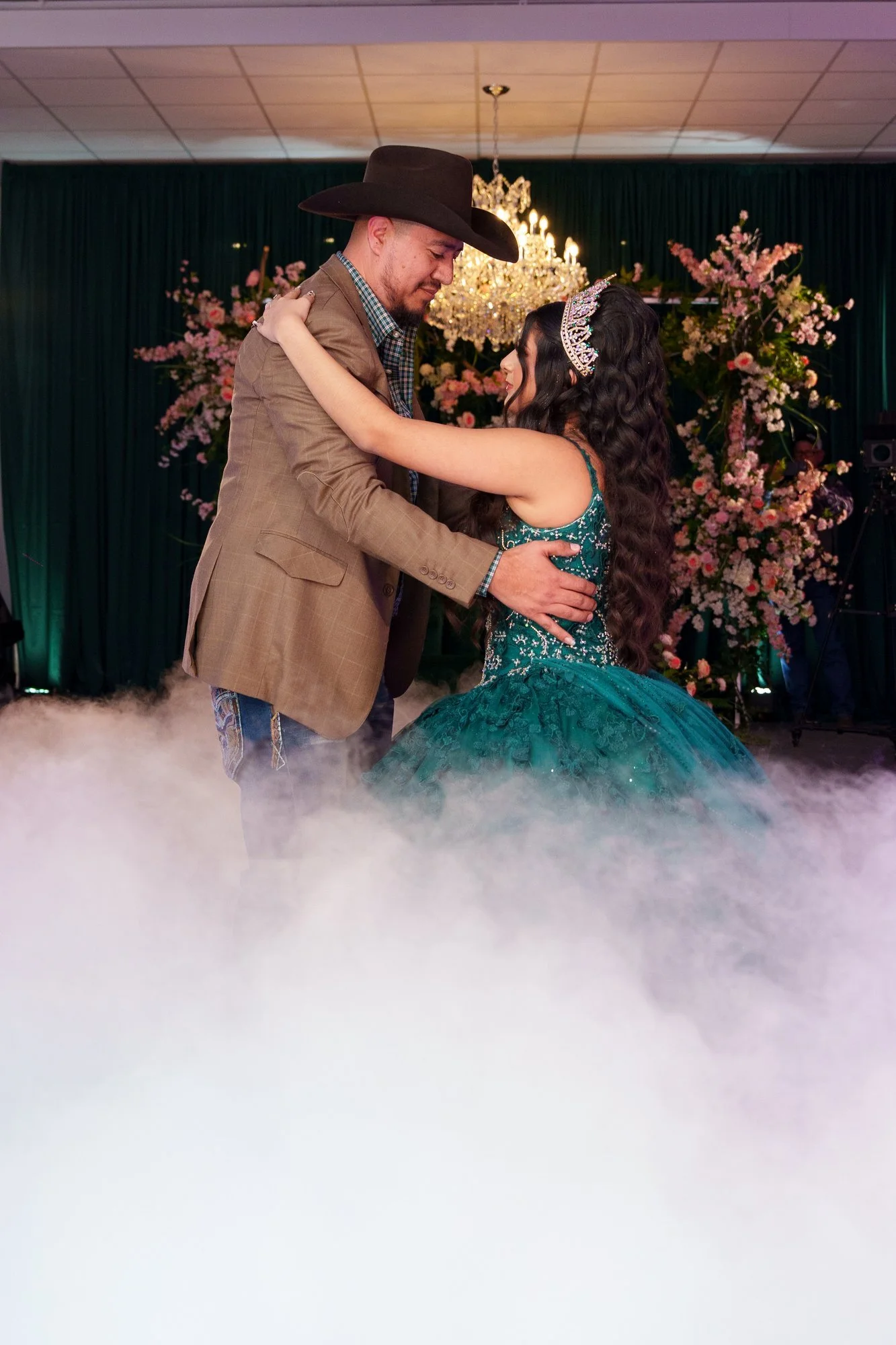 Nevaeh embracing her father in a cowboy hat during the father-daughter dance, low-lying fog, chandelier and pink florals in the background