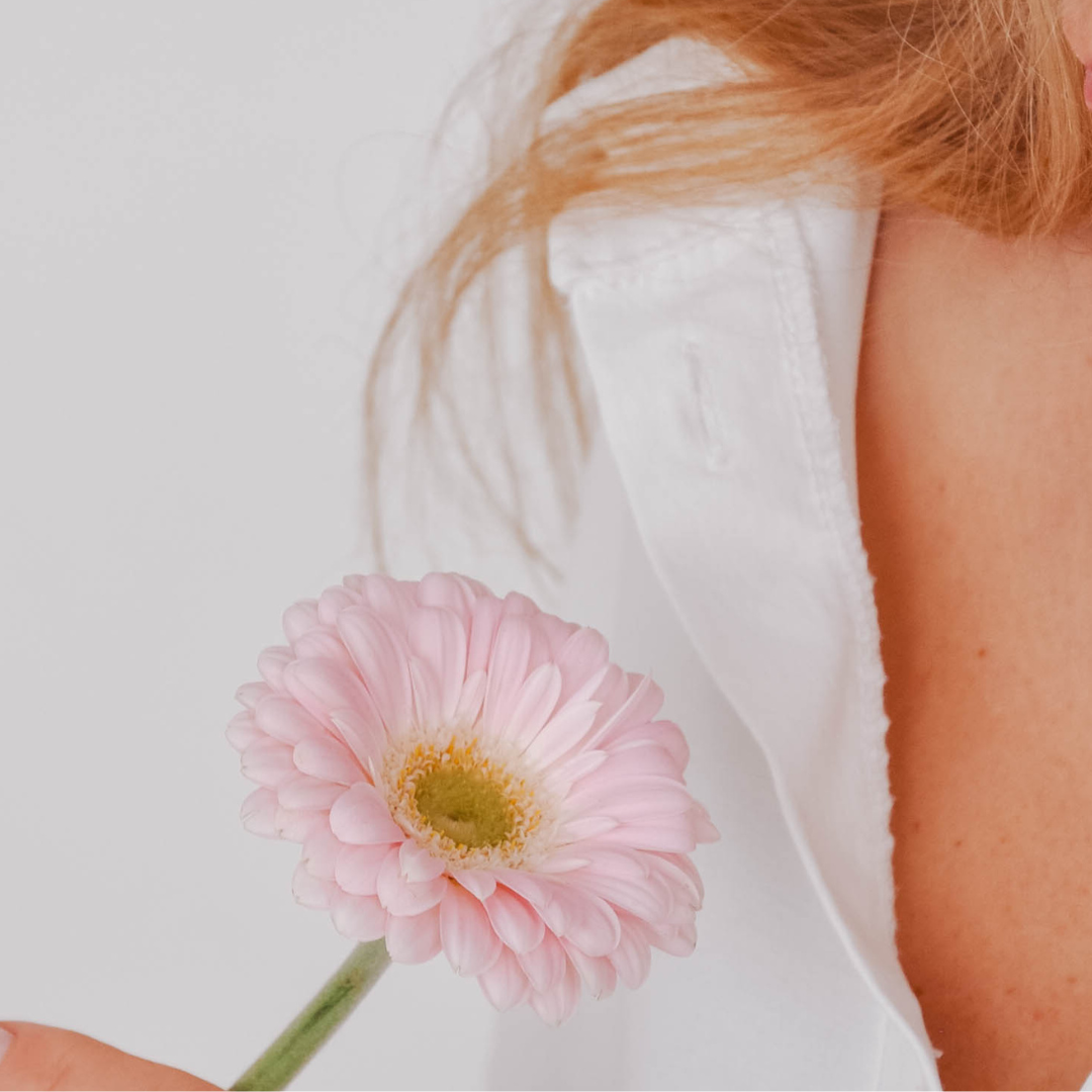 Close-up of a woman holding a light pink daisy flower, wearing a white sleeveless top against a plain background.