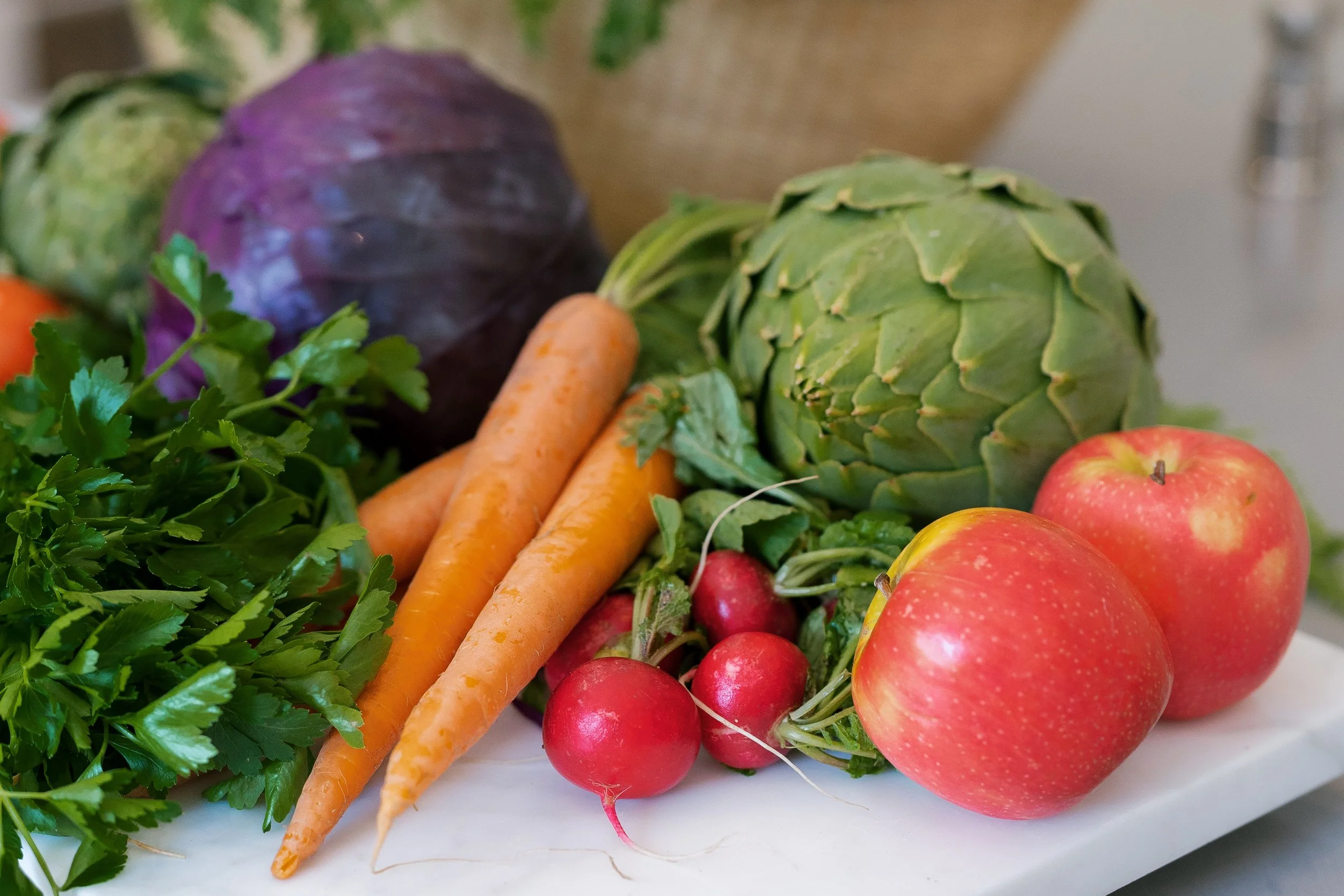 Fresh vegetables including carrots, radishes, an artichoke, apples, lettuce, purple cabbage, and vegetables in the background.