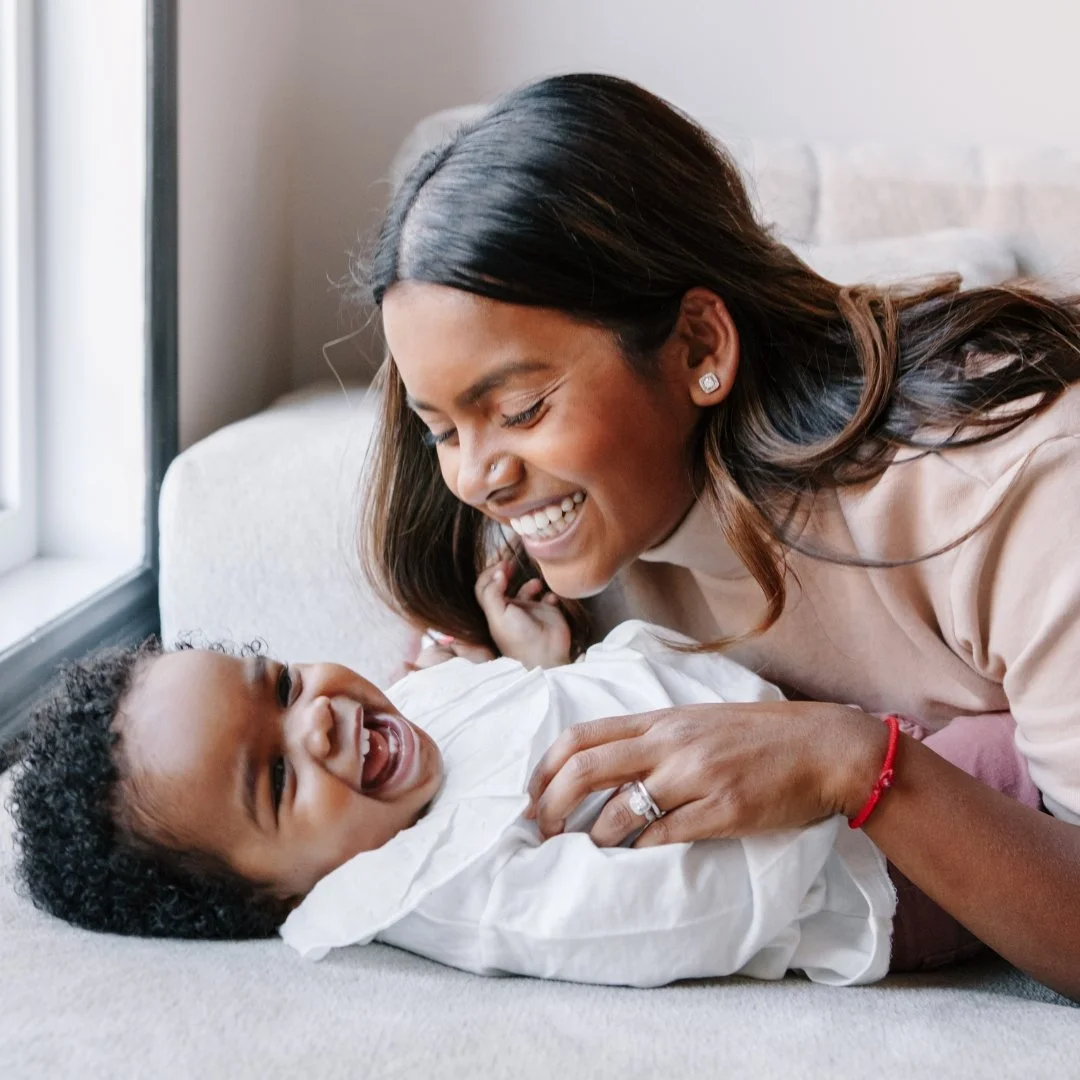 A woman and a young boy smiling and playing together on a couch indoors near a window.