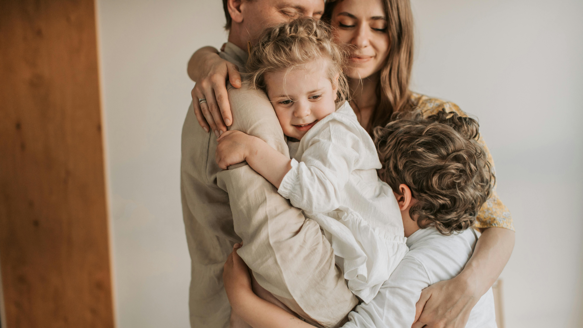 Family embracing hugs in a close, warm hug indoors, smiling and showing affection.