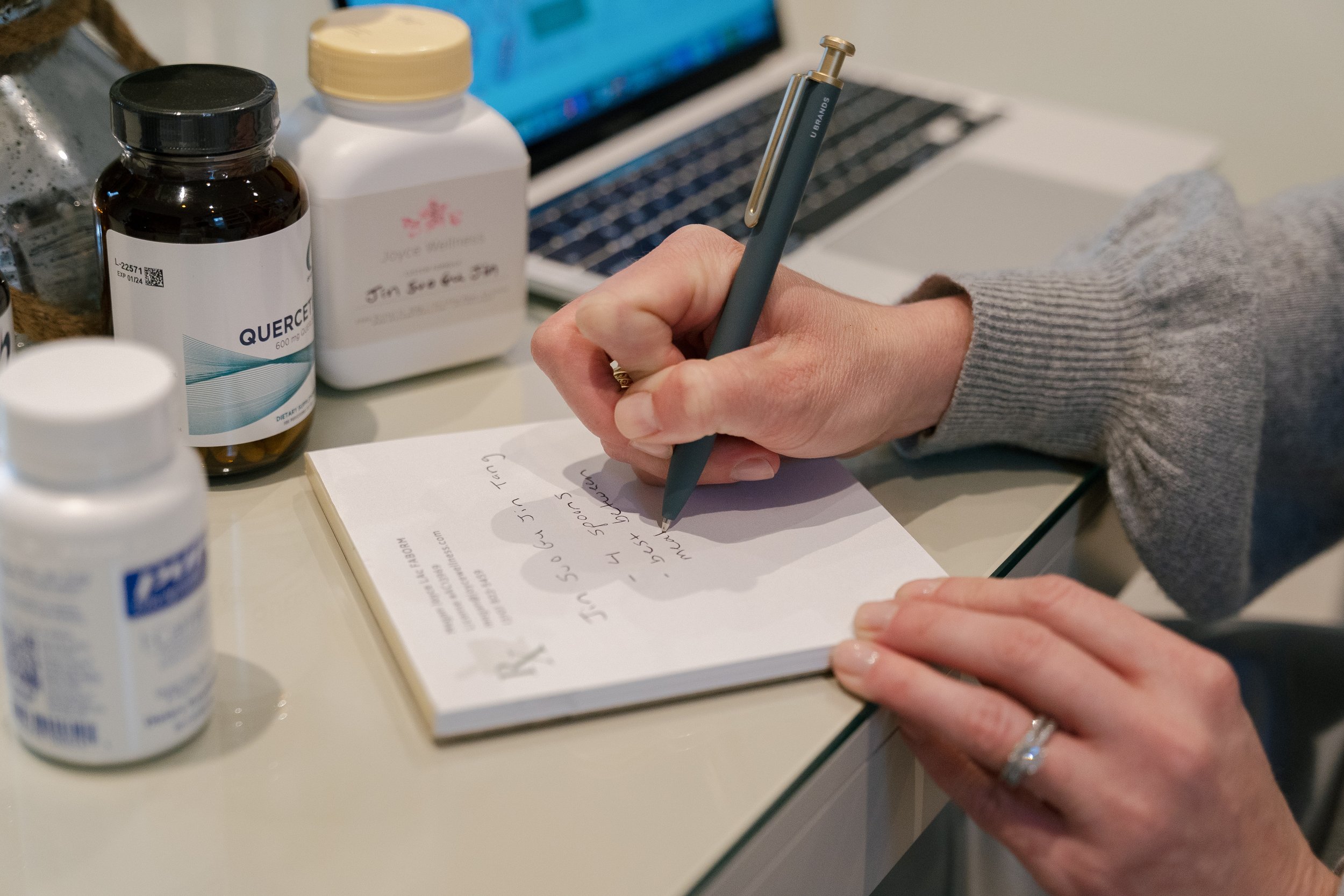A person writing in a prescription notebook with a green pen, surrounded by medication bottles on a white table.