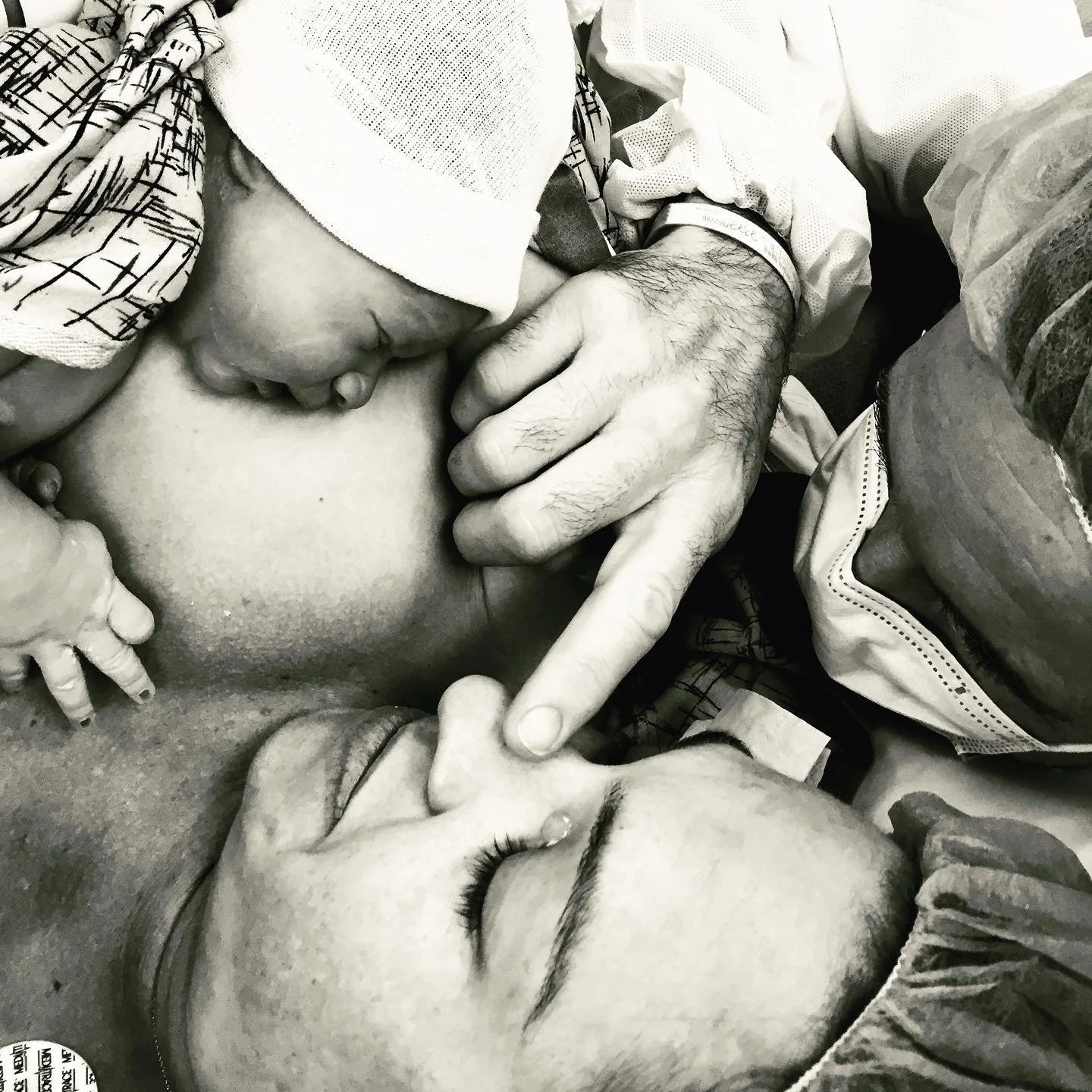 A woman and a man are holding hands and a newborn baby. The woman, wearing a hair cover, has her eyes closed with a tear rolling down her cheek.